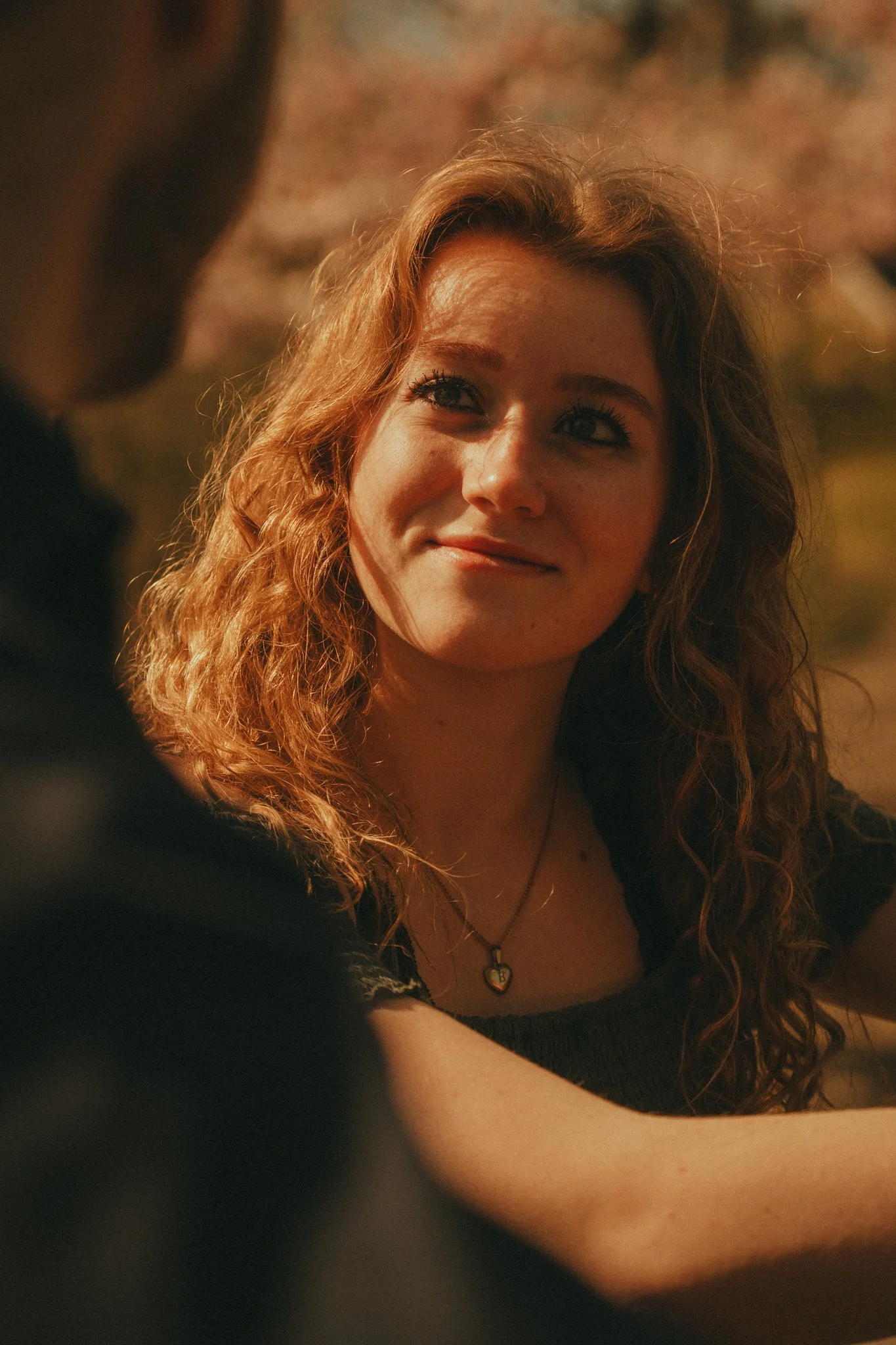 A young woman with curly red hair is smiling at someone outside the frame, with warm lighting highlighting her face and hair.