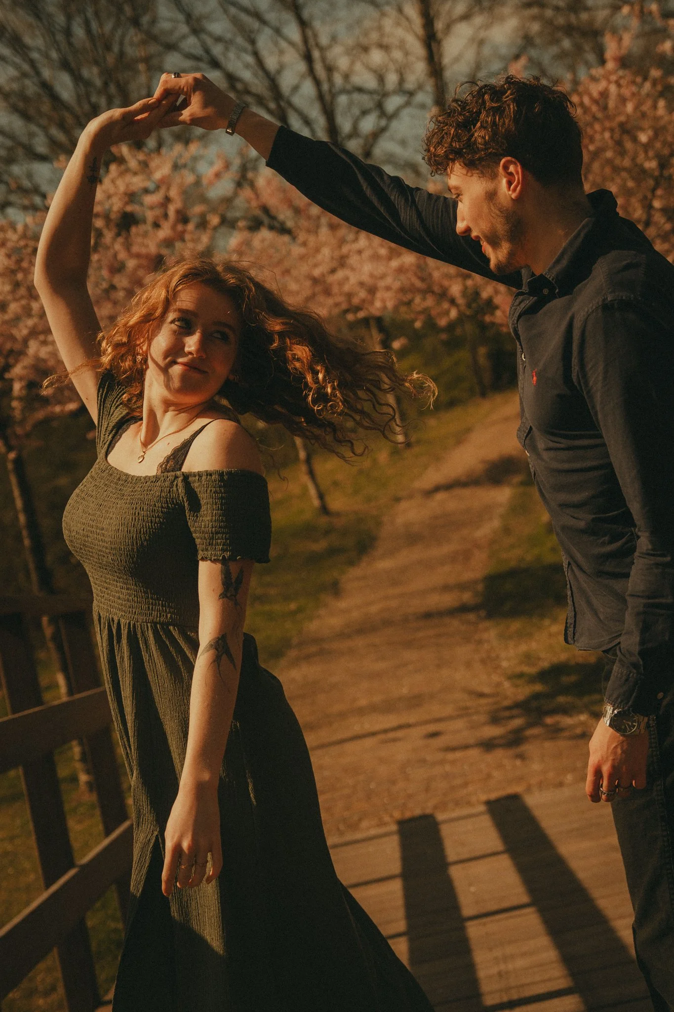 A man and woman dancing outdoors at night, holding hands with a backdrop of pink cherry blossom trees and a wooden walkway, soft warm lighting.
