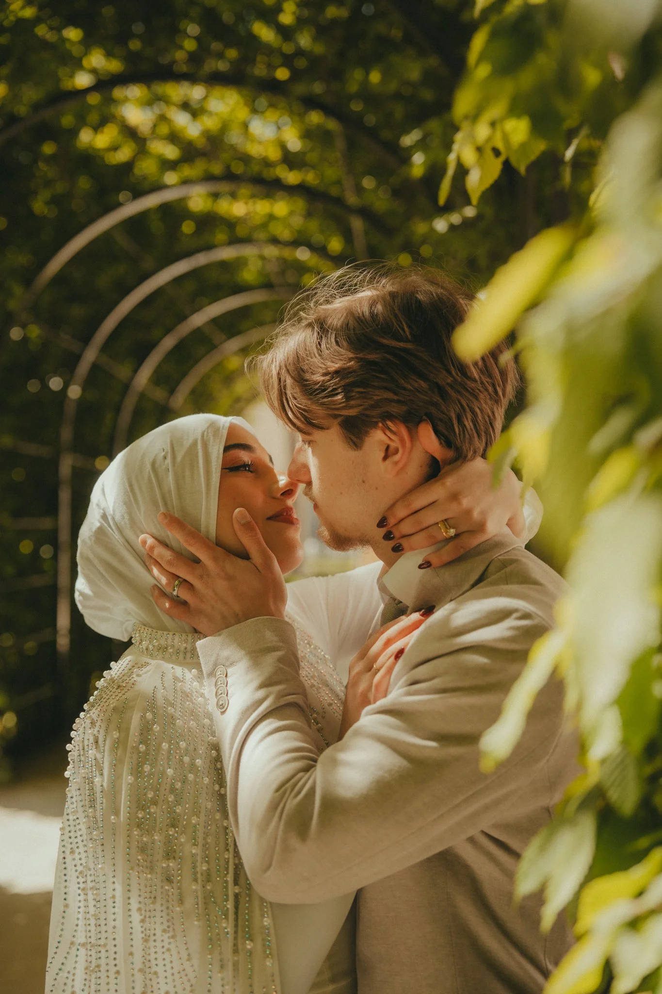 A couple is kissing, with the woman wearing a white hijab and the man in a beige suit. They are holding each other tenderly, surrounded by green foliage, illuminated by soft lighting at night.