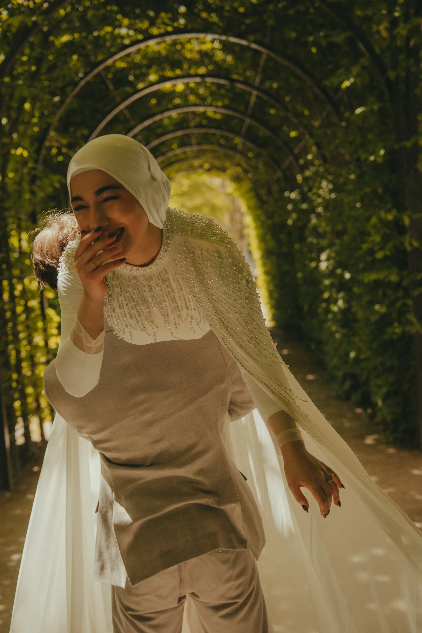A woman wearing a beige dress and a white headscarf, seemingly laughing and covering her mouth, with a green leafy tunnel in the background.