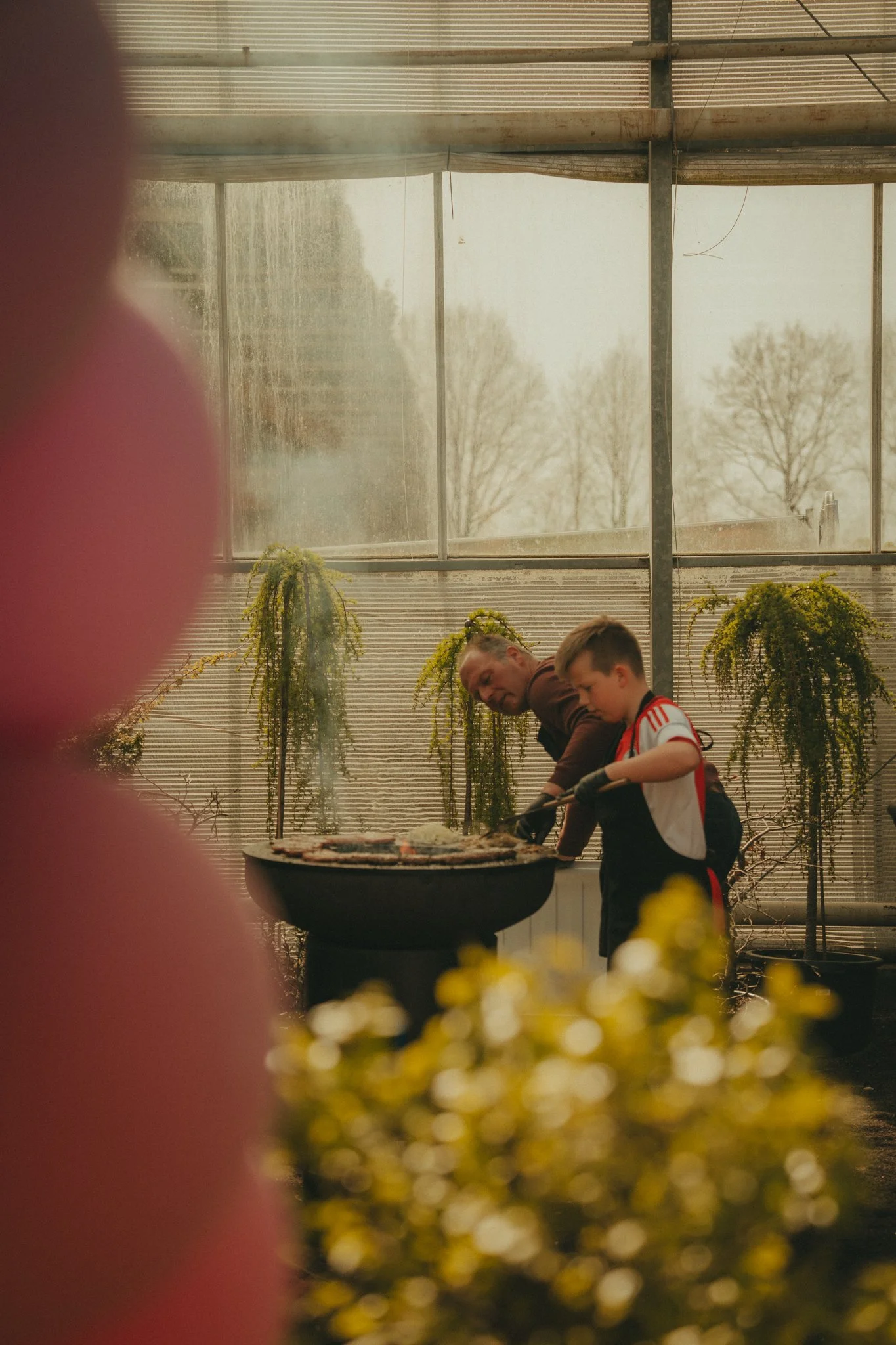 Two people, a man and a boy, are standing by a barbecue grill inside a greenhouse with hanging plants and large windows showing trees outside.