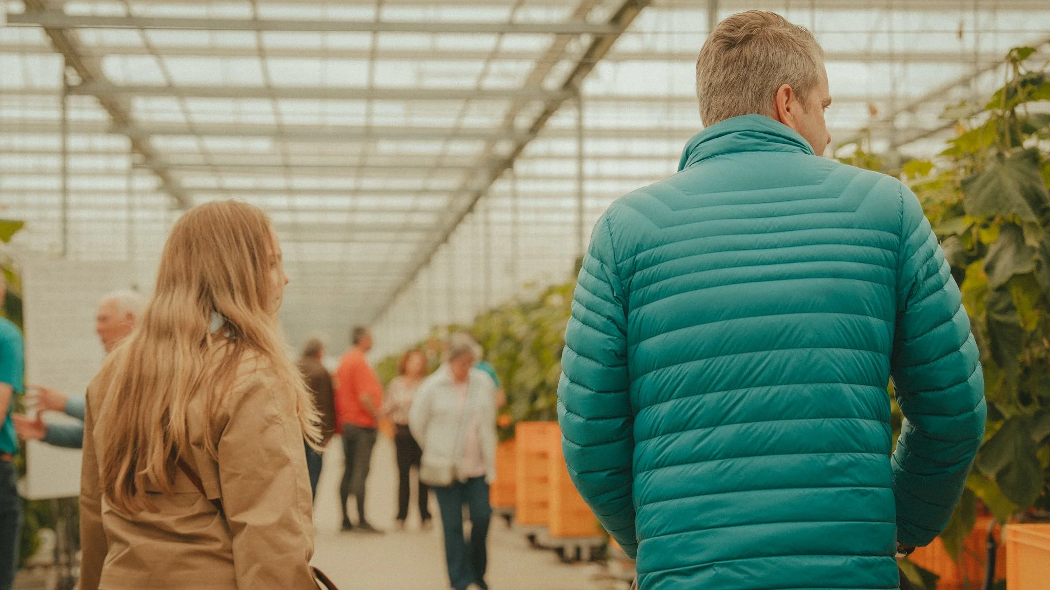 A woman with long blonde hair and a man with short blonde hair are standing inside a greenhouse, observing plants. Multiple people are in the background, some talking and walking among the plants.
