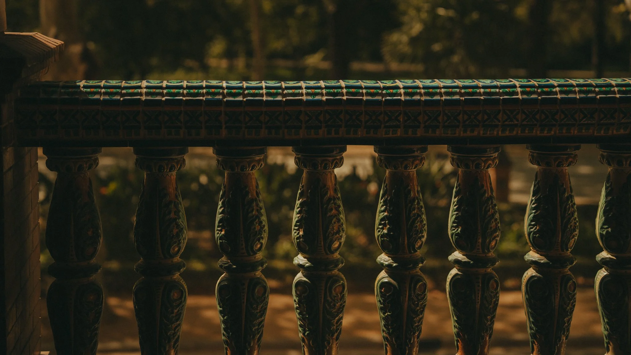 Ornate wooden balcony railing with colorful ceramic tiles on top, viewed during sunset or dusk with blurred trees in the background.