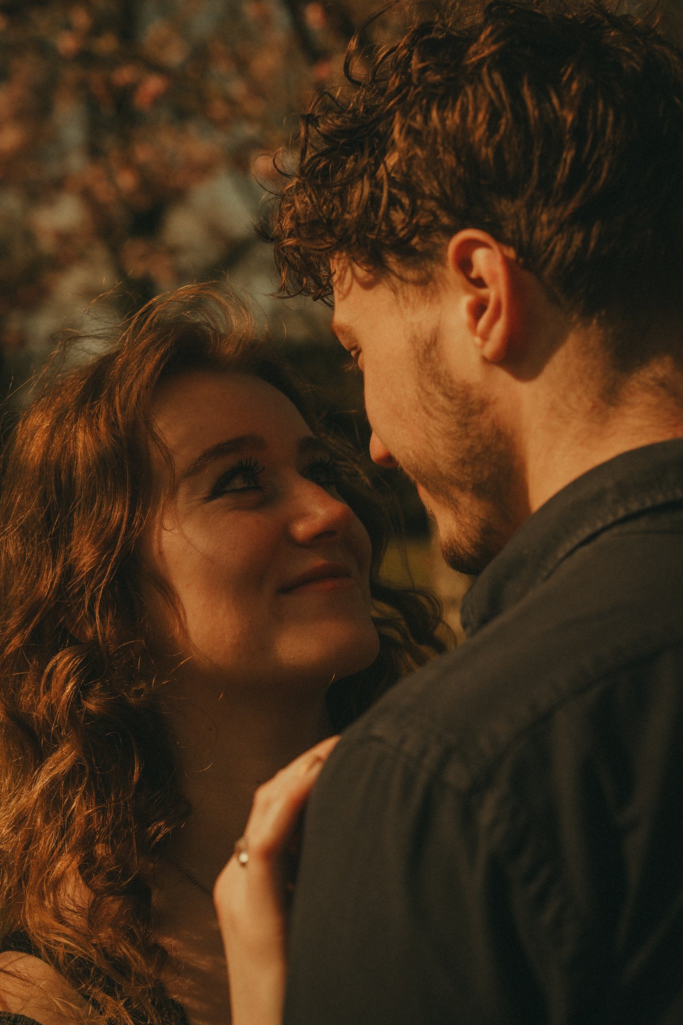 A young couple close together, gazing into each other's eyes, with the woman's face and the man's profile visible, outdoors during sunset.