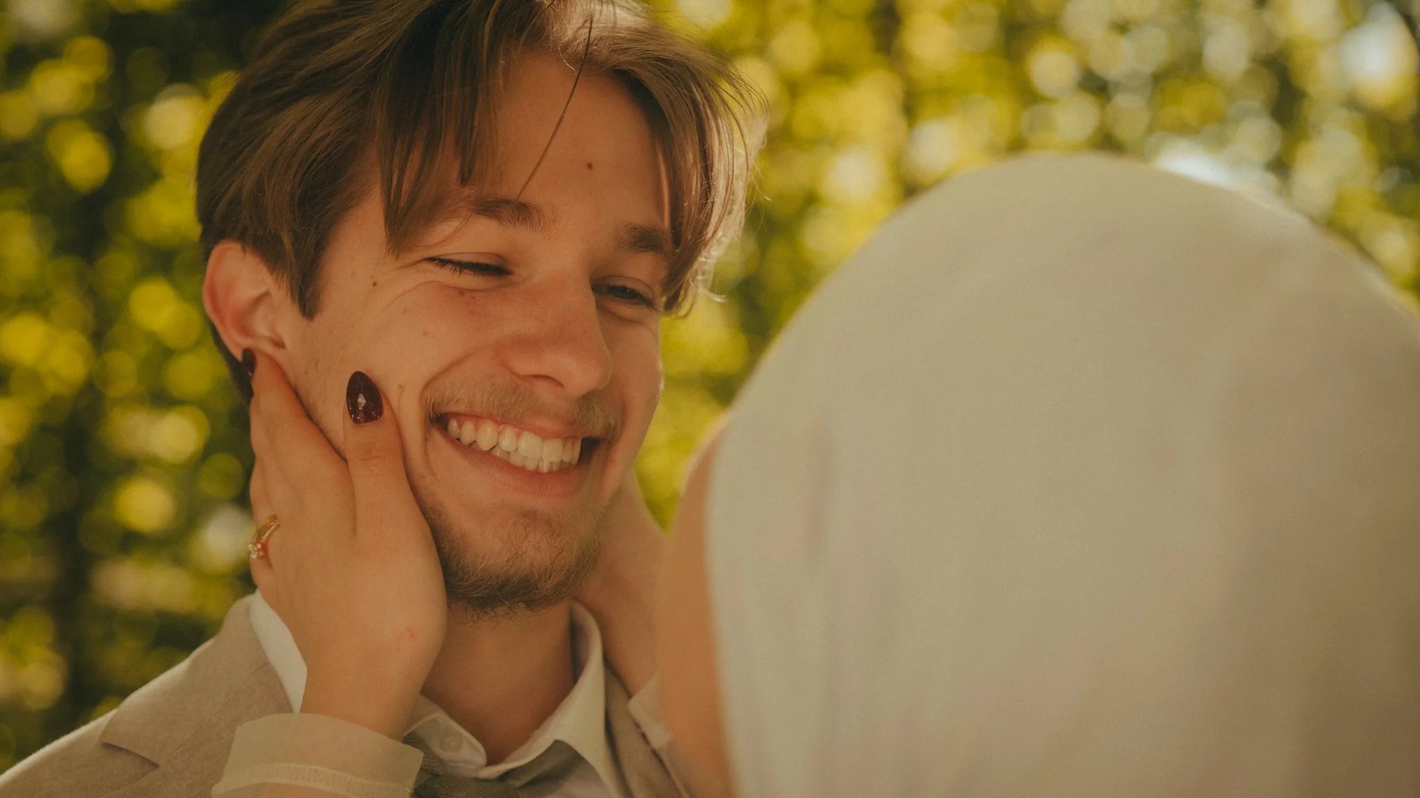 A happy man with brown hair and a beard, smiling while holding the sides of his head, outdoors with blurred yellow and green background.