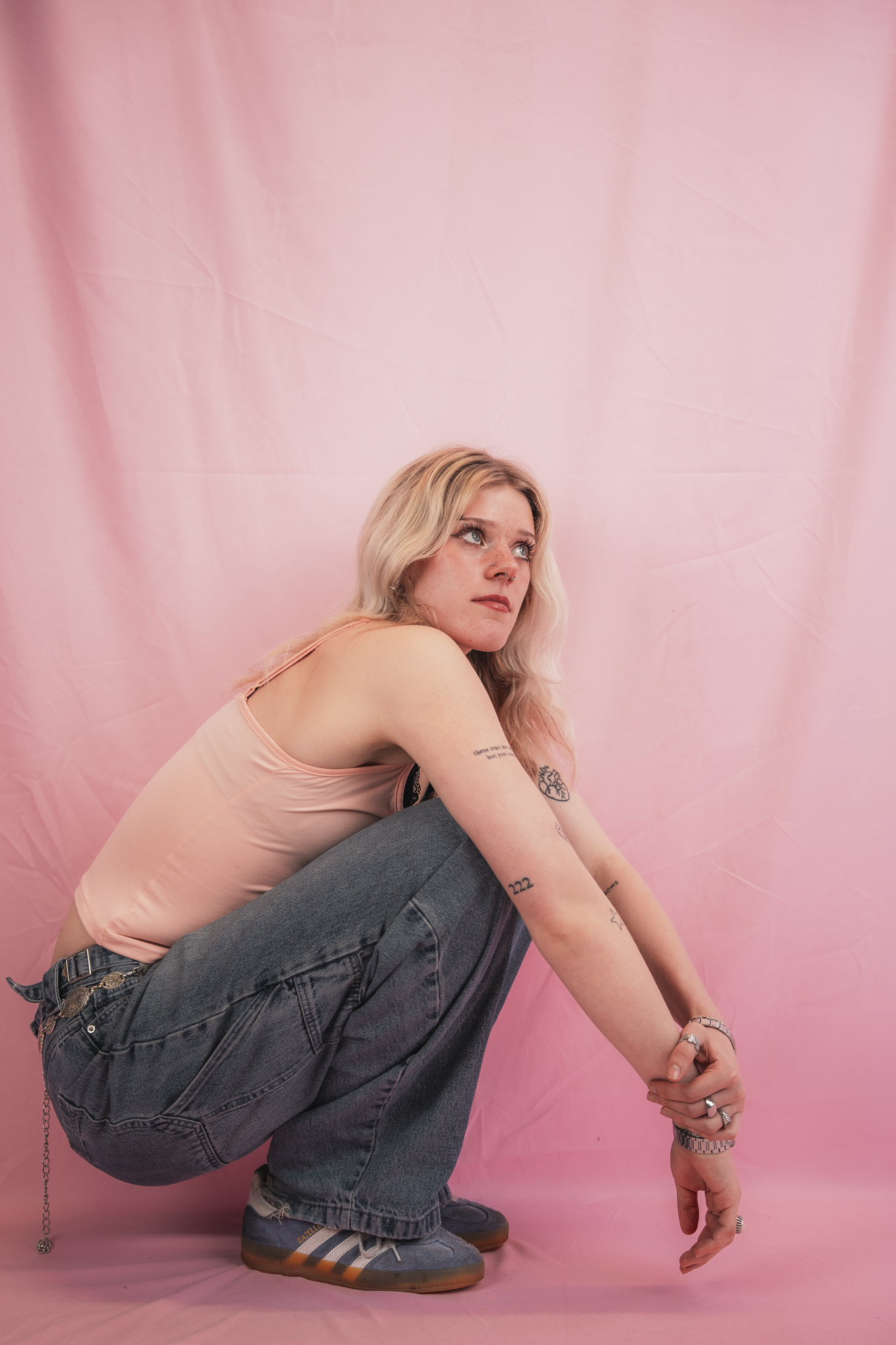 A person with blonde hair, tattoos, and rings on their fingers, squatting against a pink backdrop, wearing a light pink tank top, gray jeans, and Adidas sneakers.