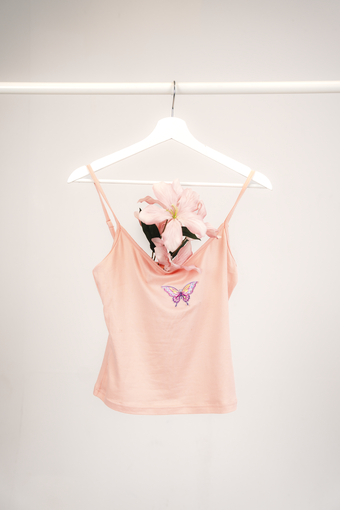 Pink satin camisole with butterfly embroidery hanging on a white hanger from a clothing rod with a pink flower inside.