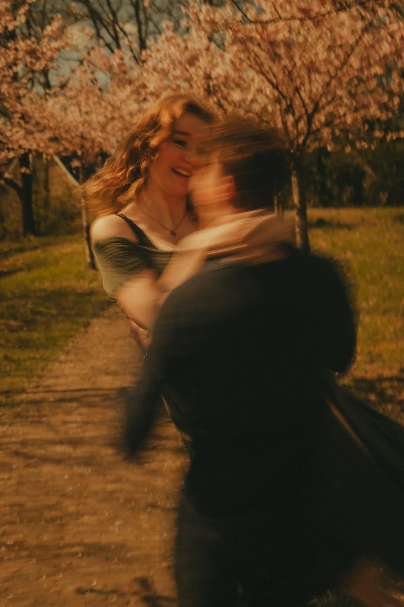 A young woman with curly hair smiling and hugging a person outdoors at night, with pink flowering trees in the background.