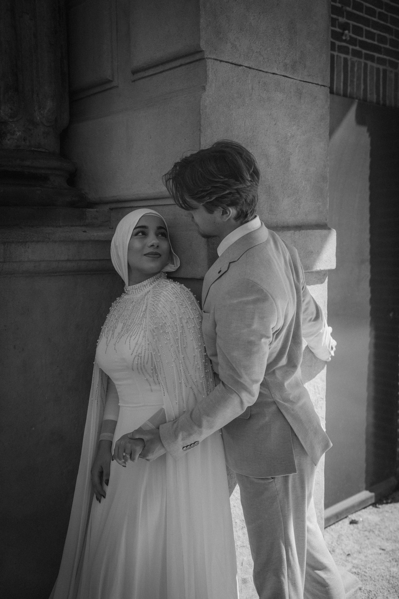 A black-and-white photo of a man and woman standing close together outside, leaning against a stone building wall. The woman is wearing a hijab and a beaded dress, while the man is dressed in a light-colored suit. They are gazing into each other's ey