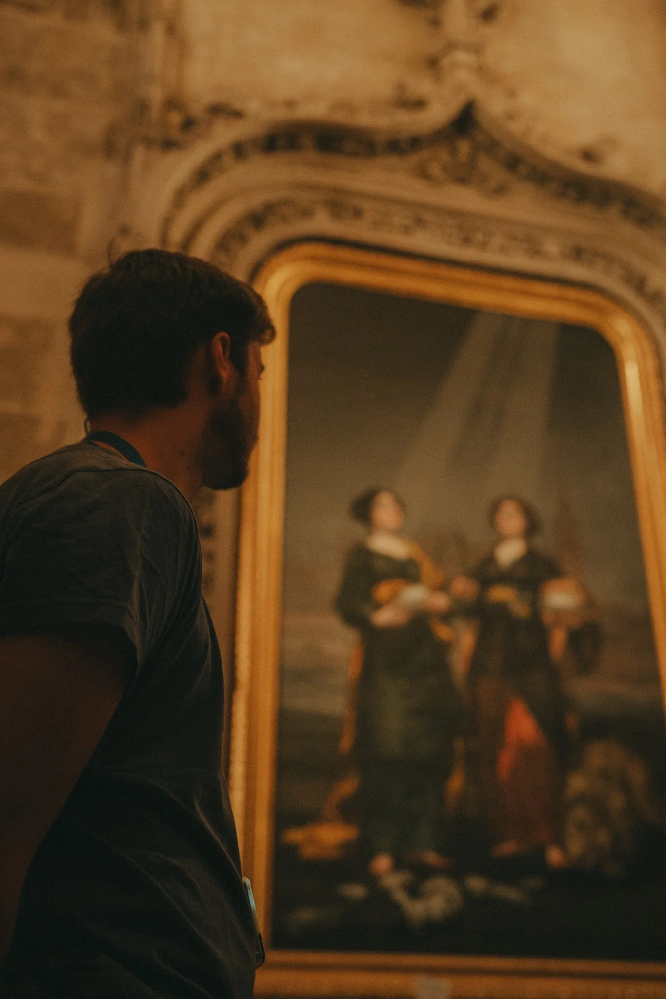 A man with brown hair and a beard looking at a large framed painting of two men playing guitars in a gallery with ornate ceiling details.