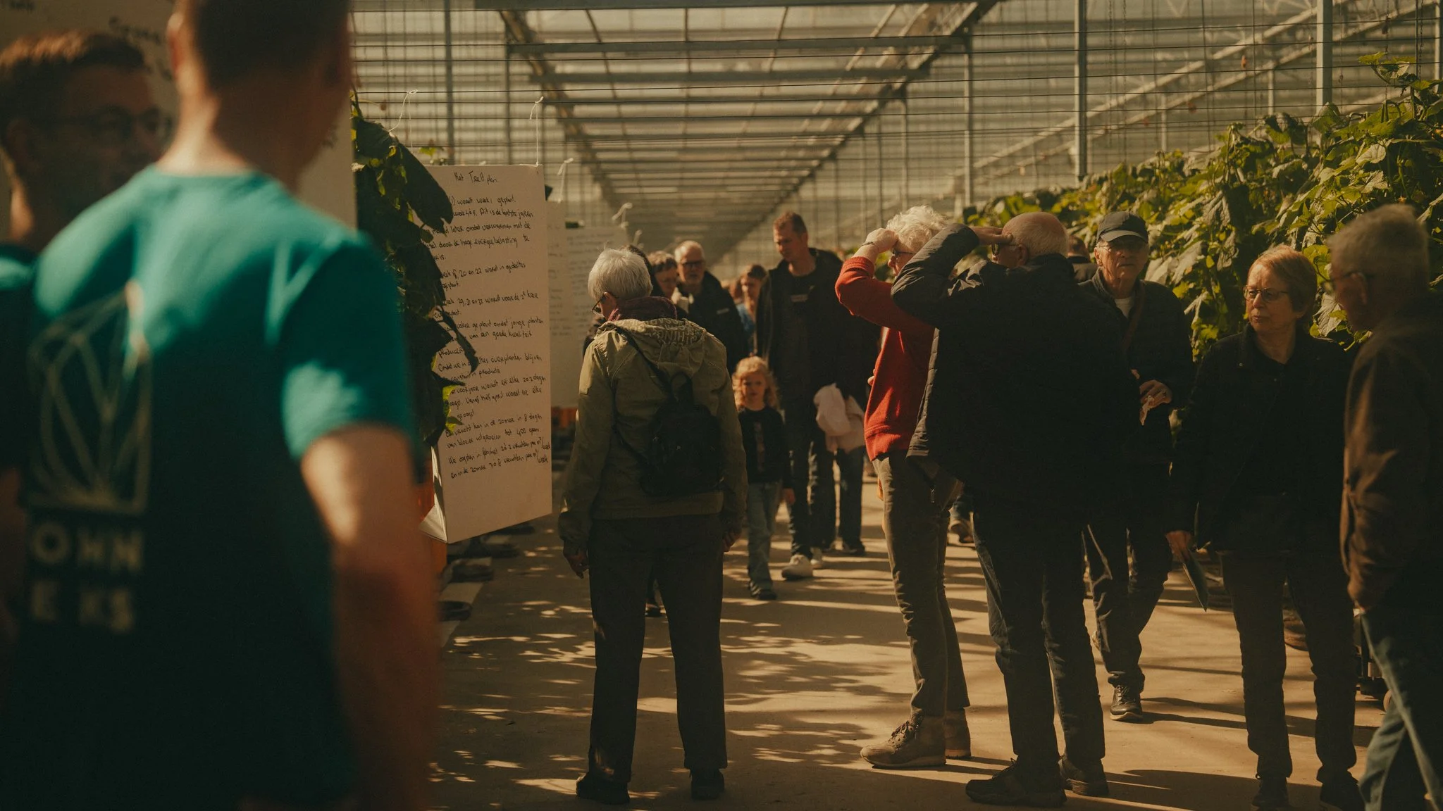 People gathered inside a greenhouse or plant nursery, engaging in conversations and observing plants and information boards.