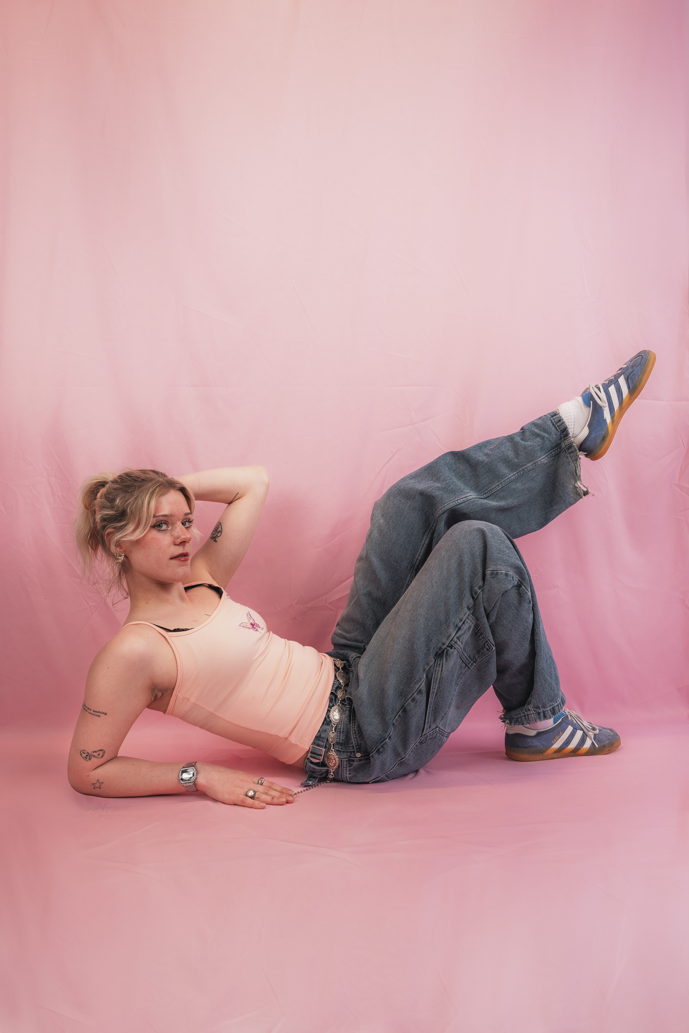 A young woman lying on the floor with her legs raised, wearing a light pink tank top, dark jeans, and sneakers, against a pink background.