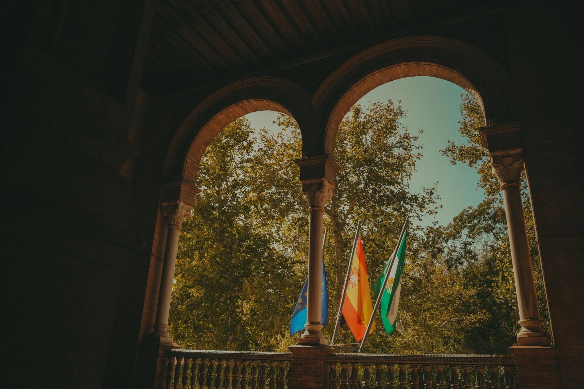 View of an outdoor archway with trees and sky visible through the arches, and colorful flags hanging on poles below.