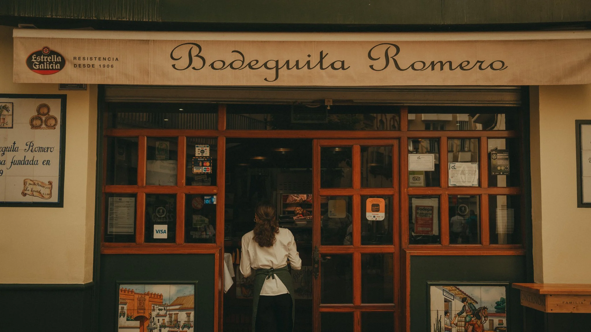 The storefront of Bodega Romero with a beige awning, wood-framed glass windows, and a person with long hair and a white shirt standing in front.