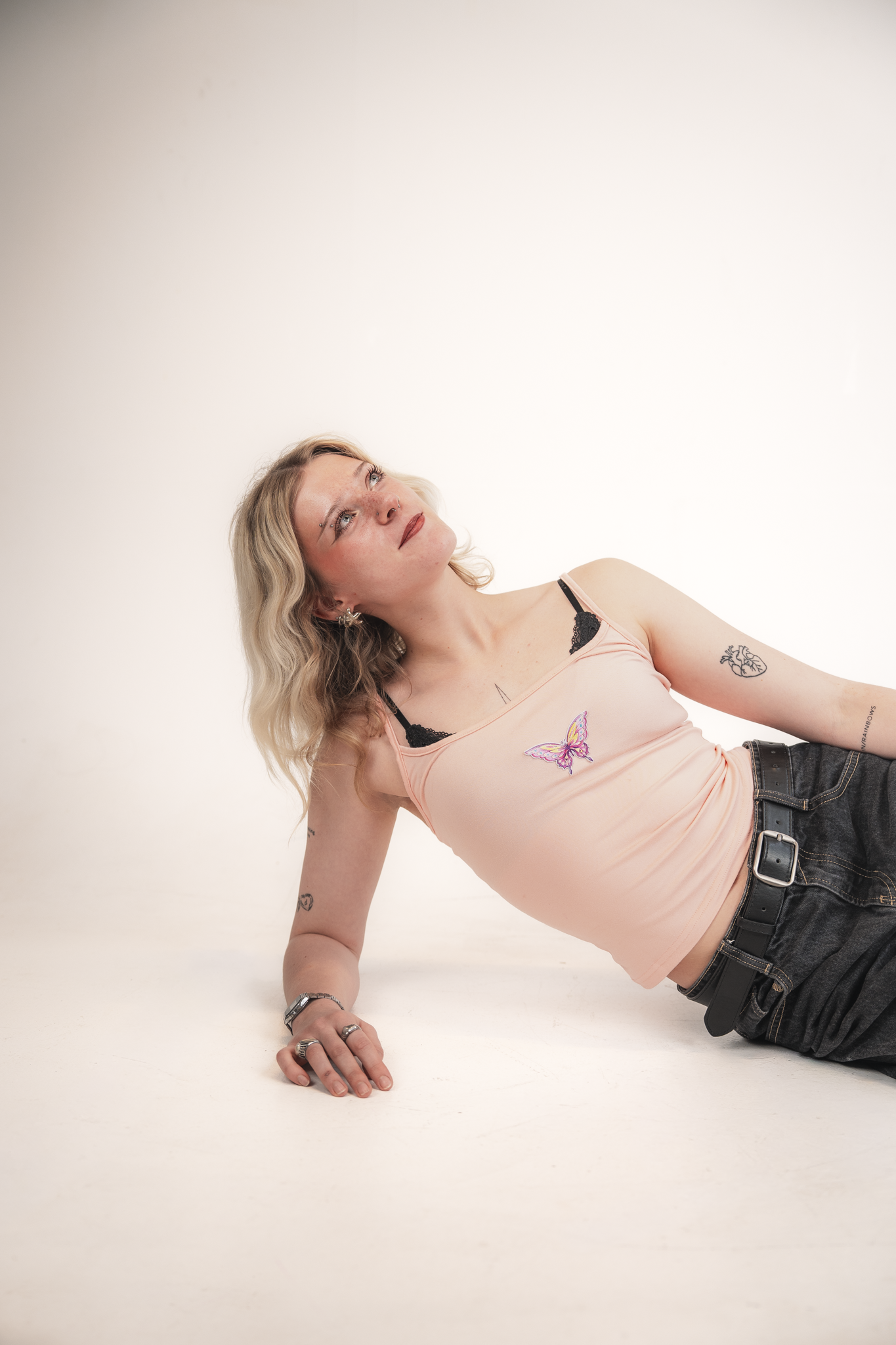 A woman with blonde hair, wearing a pink tank top with a butterfly design, black jeans, and multiple rings, is lying on her side on a white background. She is looking upward with a thoughtful expression.