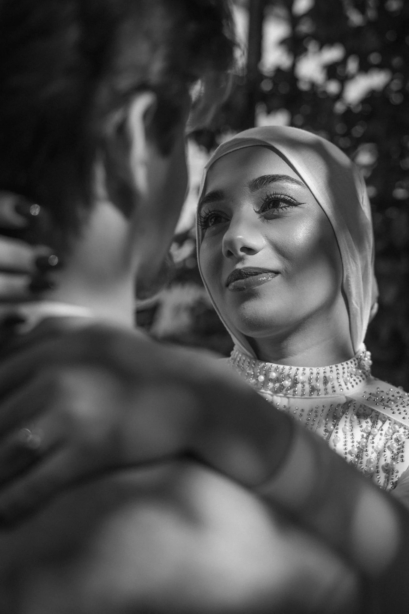 A woman wearing a headscarf and a pearl-embellished dress gazes at a man, with her face illuminated and her eyes focused on him, amid blurred background with bokeh lights.