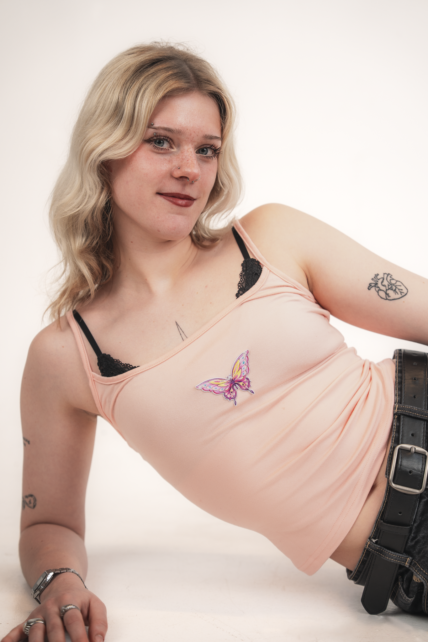 A young woman with blonde hair styled in loose waves, blue eyes, and light freckles on her face, wearing a pink spaghetti strap tank top with black lace underneath, a butterfly patch, and a tattoo of a heart with a heartbeat line on her arm. She has 