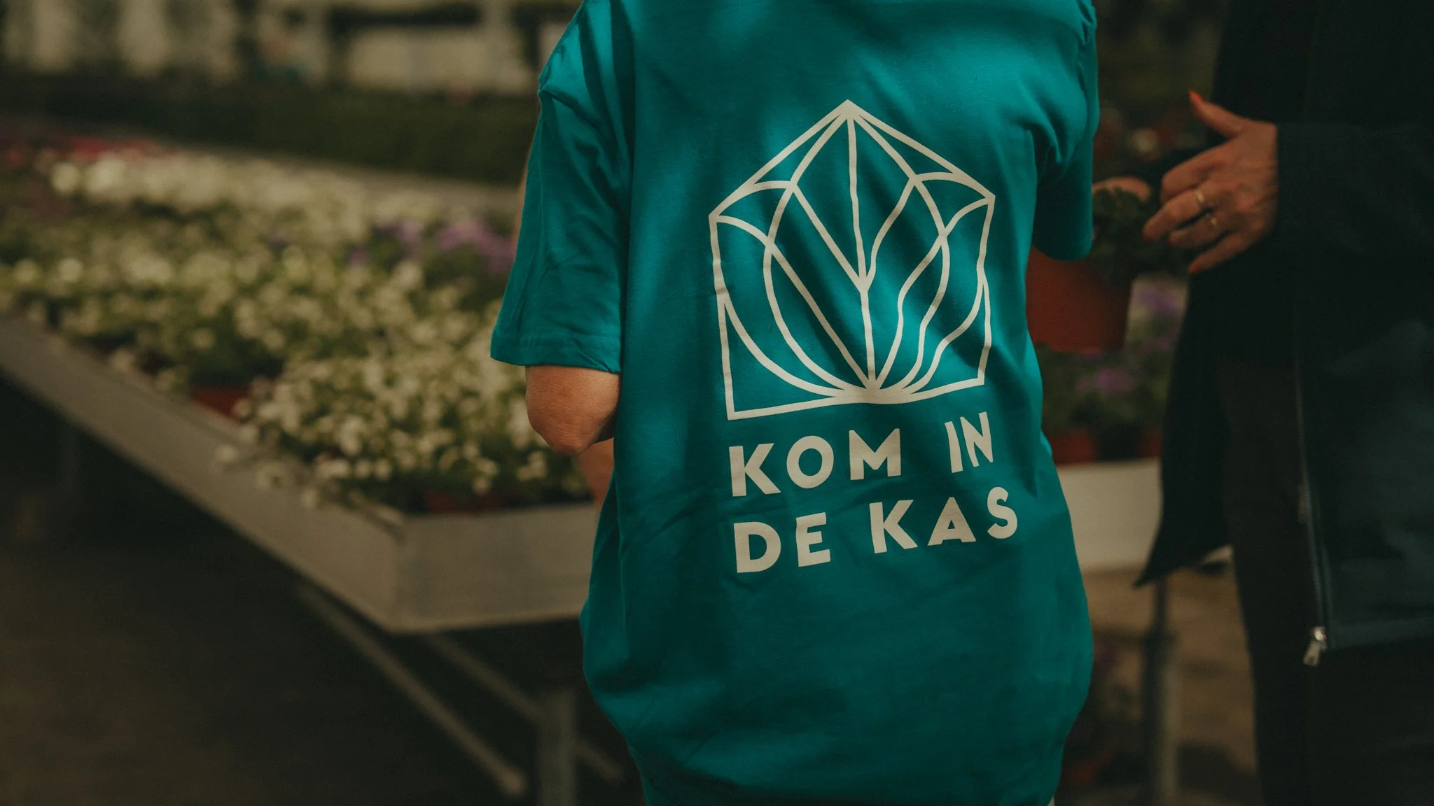 Person wearing a teal T-shirt with white geometric design and text in a non-English language, standing among potted plants in a garden store or greenhouse.