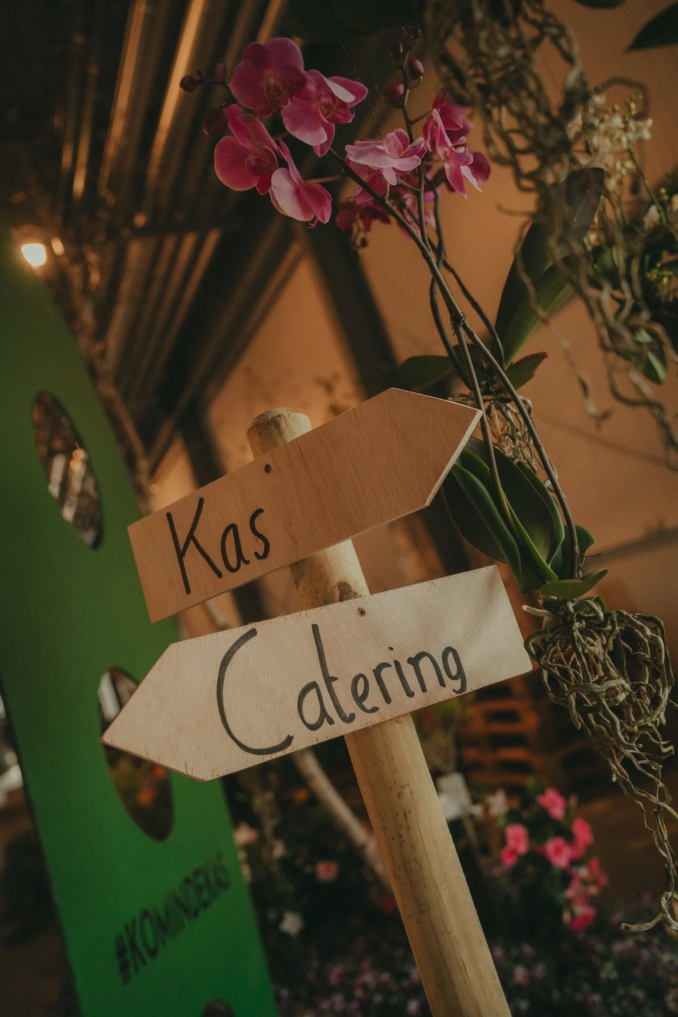 Close-up of a plant with pink orchids and a wooden plant marker with the handwritten words 'Kasi' and 'Caterina' among other signs in the background. The scene appears to be in a greenhouse or flower shop.