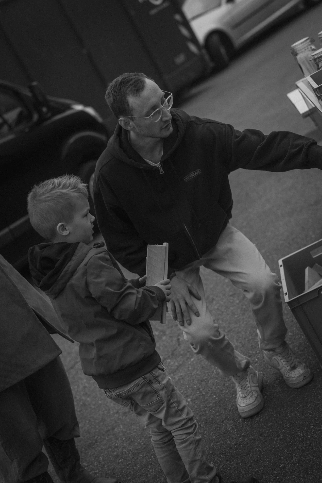 A man with glasses and wearing a hoodie is sitting on a bench talking to a young boy with blonde hair and a backpack, outdoors with cars in the background.