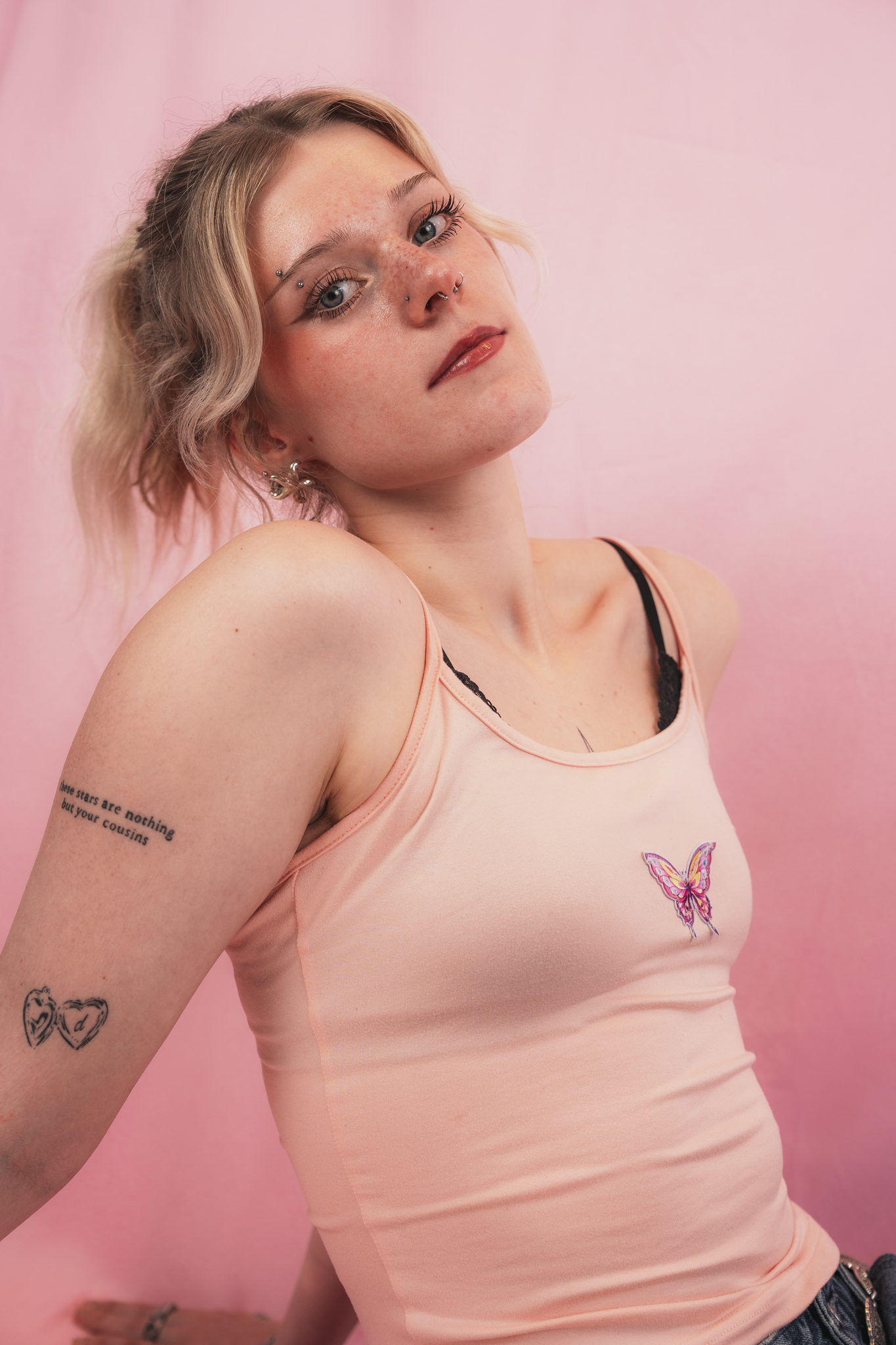 A young woman with blonde hair styled in a side part and soft waves, wearing a peach tank top with a butterfly embroidery, posing against a pink background. She has multiple facial piercings, including a nose ring, septum piercing, and eyebrow piercings. She has tattoos on her arm, including text and heart outlines. Her makeup includes mascara, eyeliner, and natural lipstick.