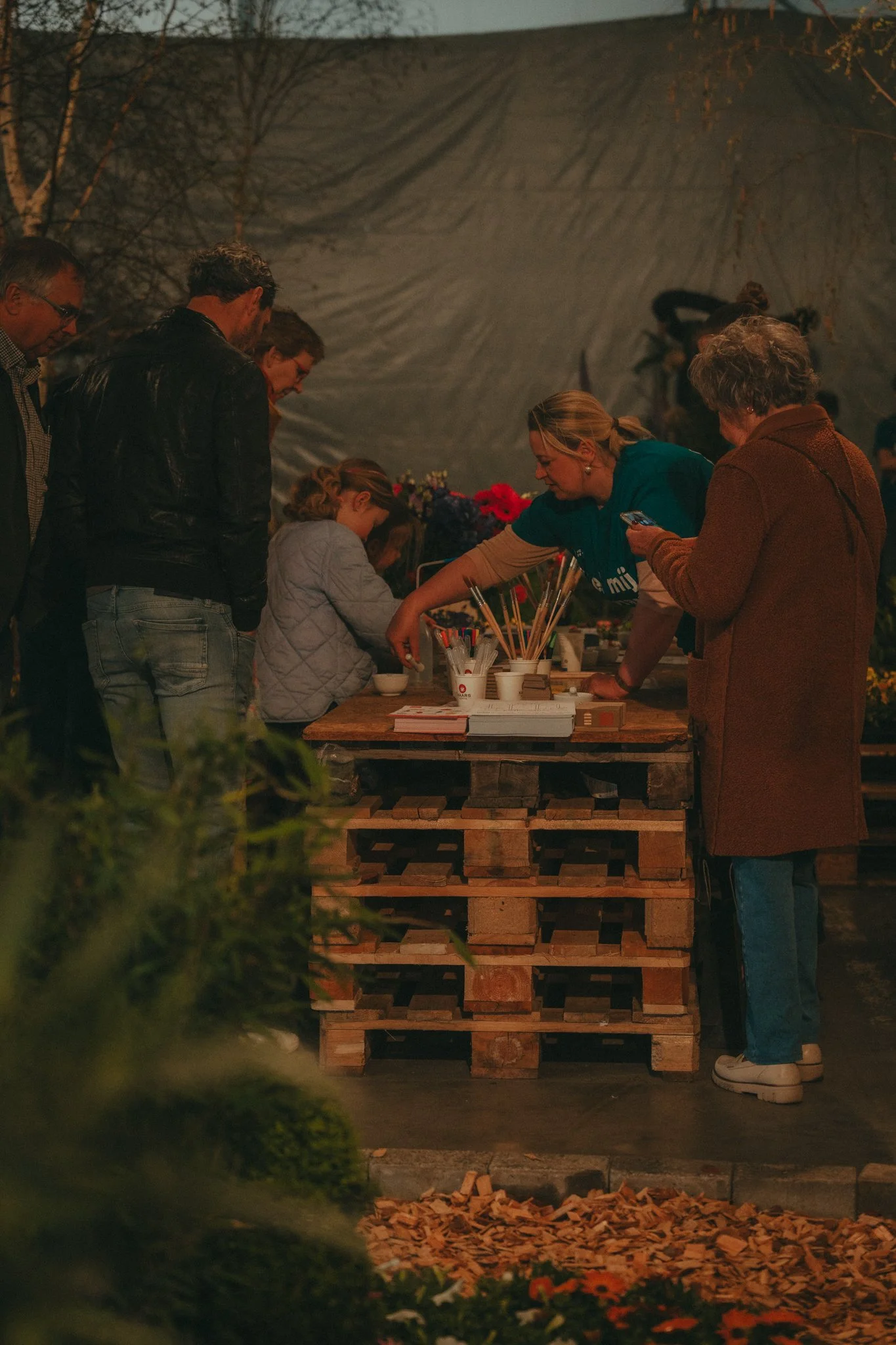People gathering around a table made of wooden pallets, with flowers and cups, outside at night.