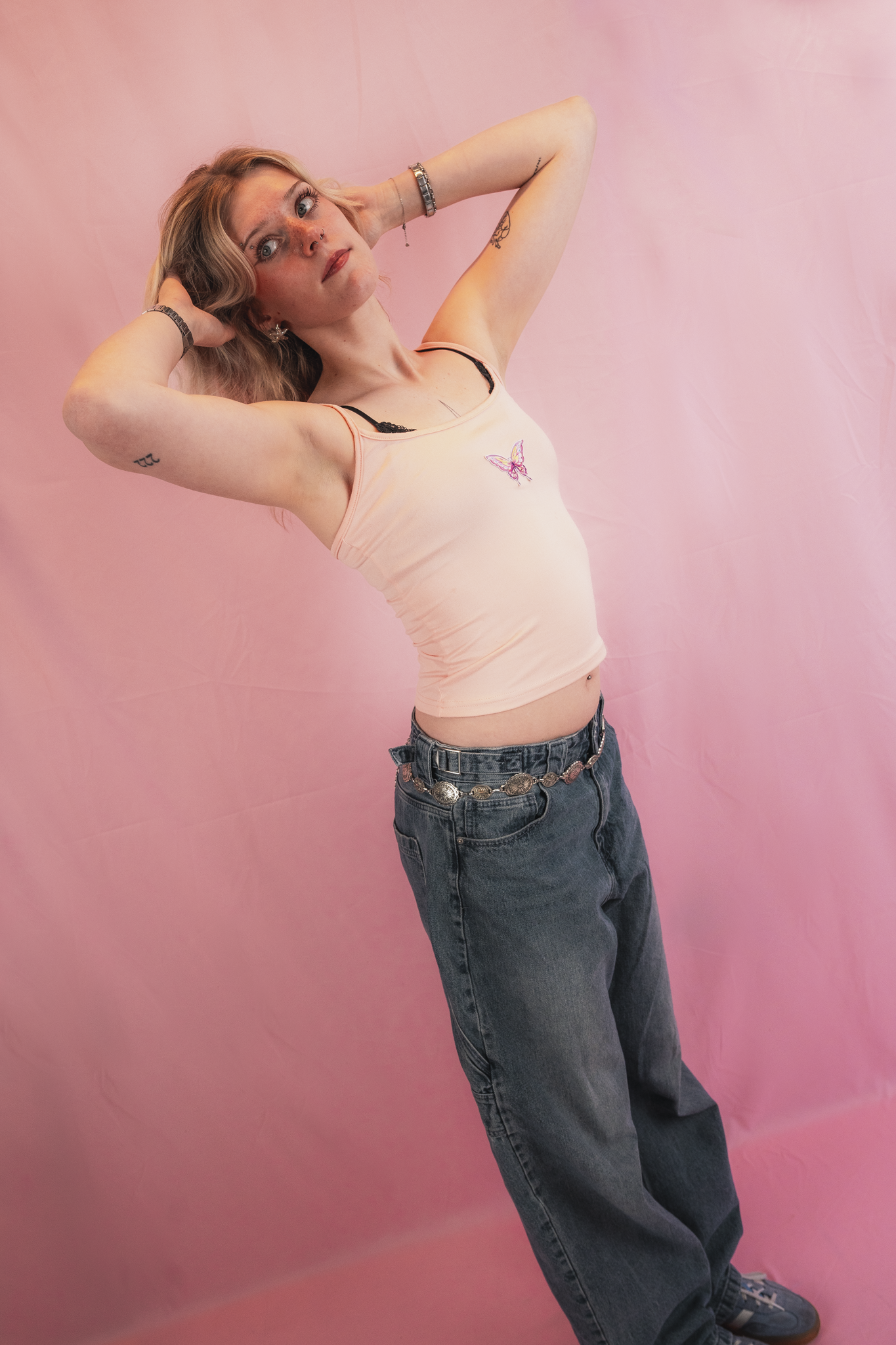 A young woman with wavy blonde hair posing against a pink backdrop, wearing a light pink tank top with a butterfly design, dark baggy jeans, and sneakers, with tattoos on her arms and a septum piercing.