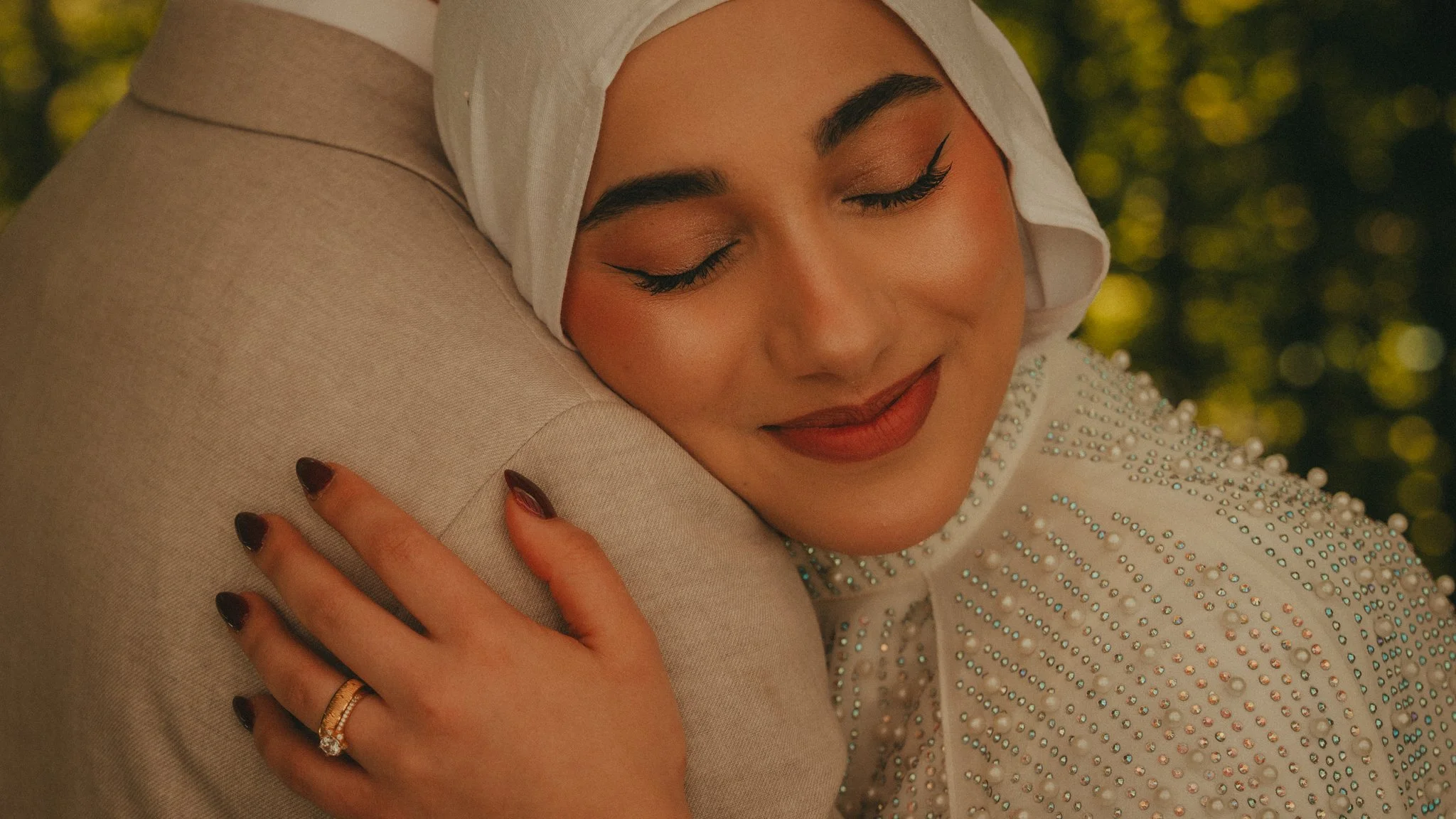 A woman with closed eyes, wearing a white headscarf and a dress decorated with beads, embraces an armrest or pillow, smiling gently. The background is blurred with yellowish-green tones.