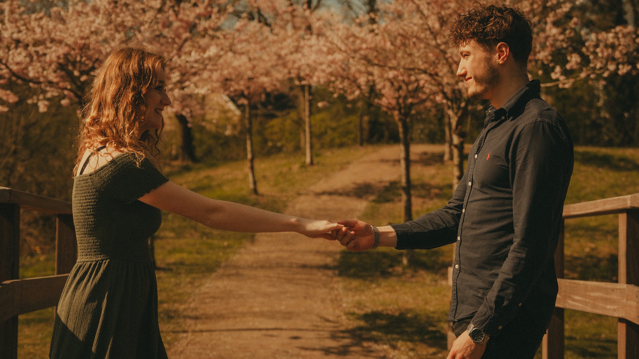 A man and woman standing outside on a wooden walkway with blooming trees in the background. They are smiling and holding hands, facing each other.