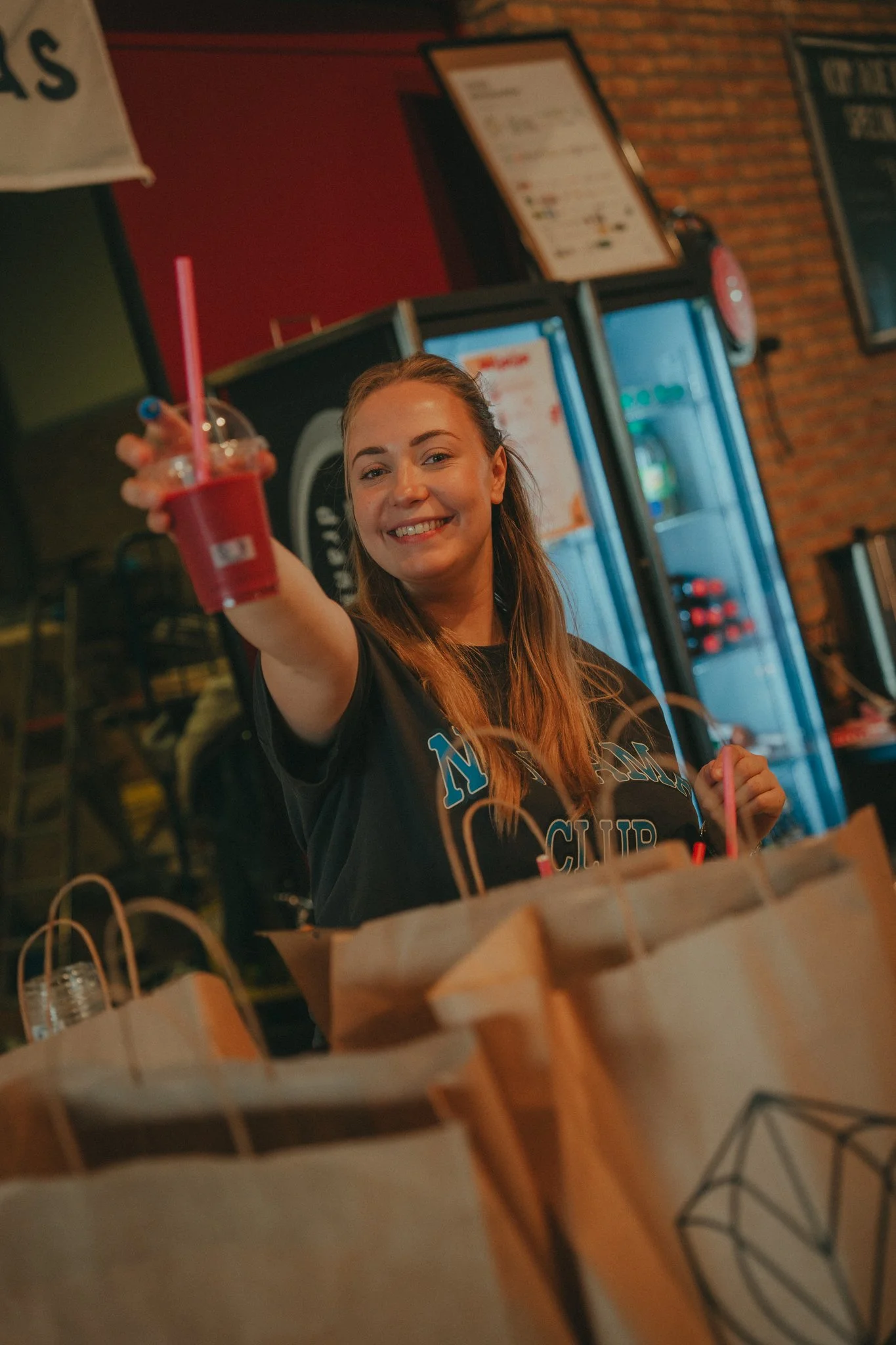 A woman with long hair smiling and holding out a pink drink in a clear cup with a straw in a bar or restaurant setting.