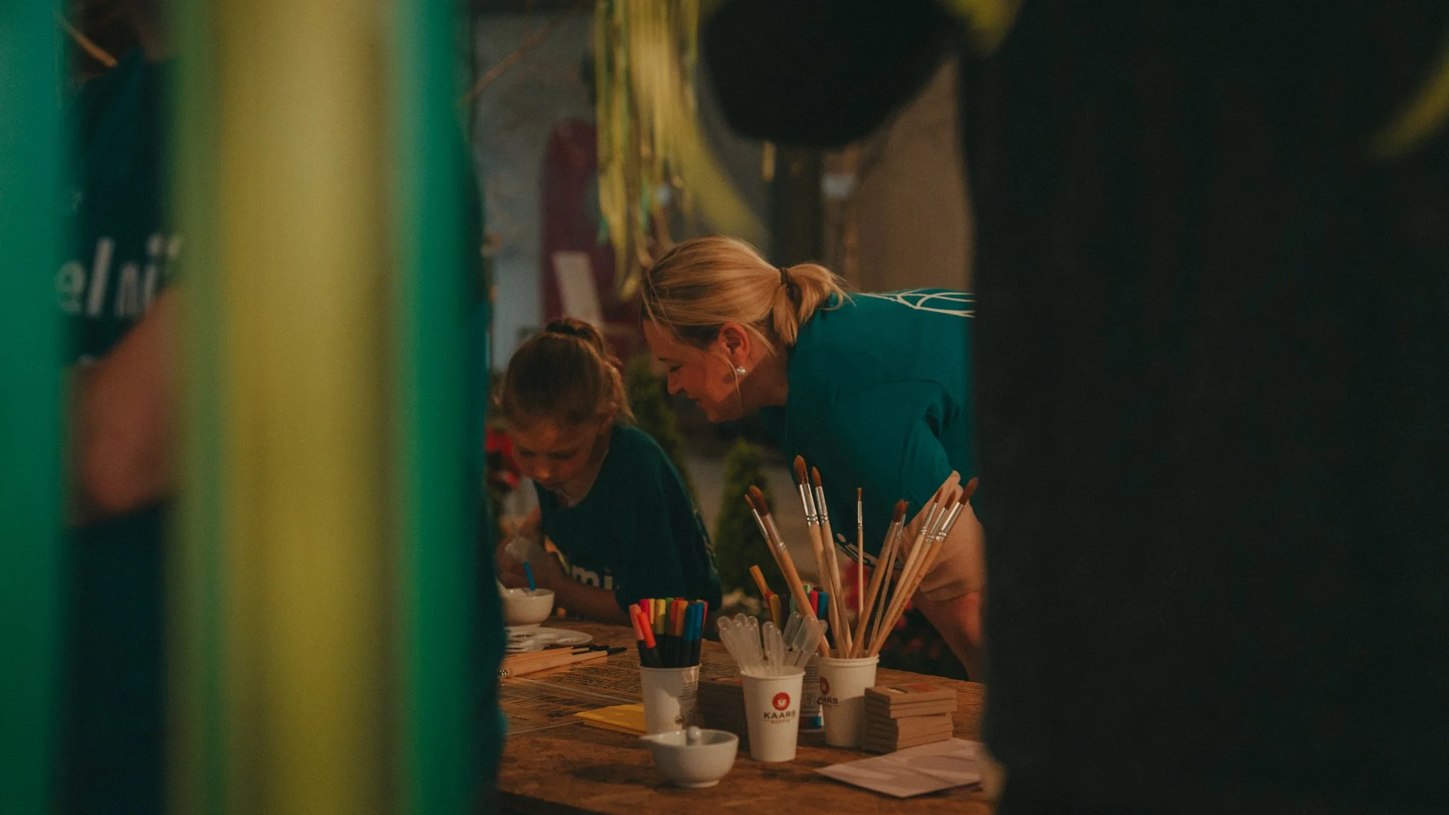 A woman with blonde hair and a young girl are working together at a table, engaging in a craft activity. The table has cups filled with colored markers and art supplies. The setting appears to be a market or craft fair, with string lights and other b