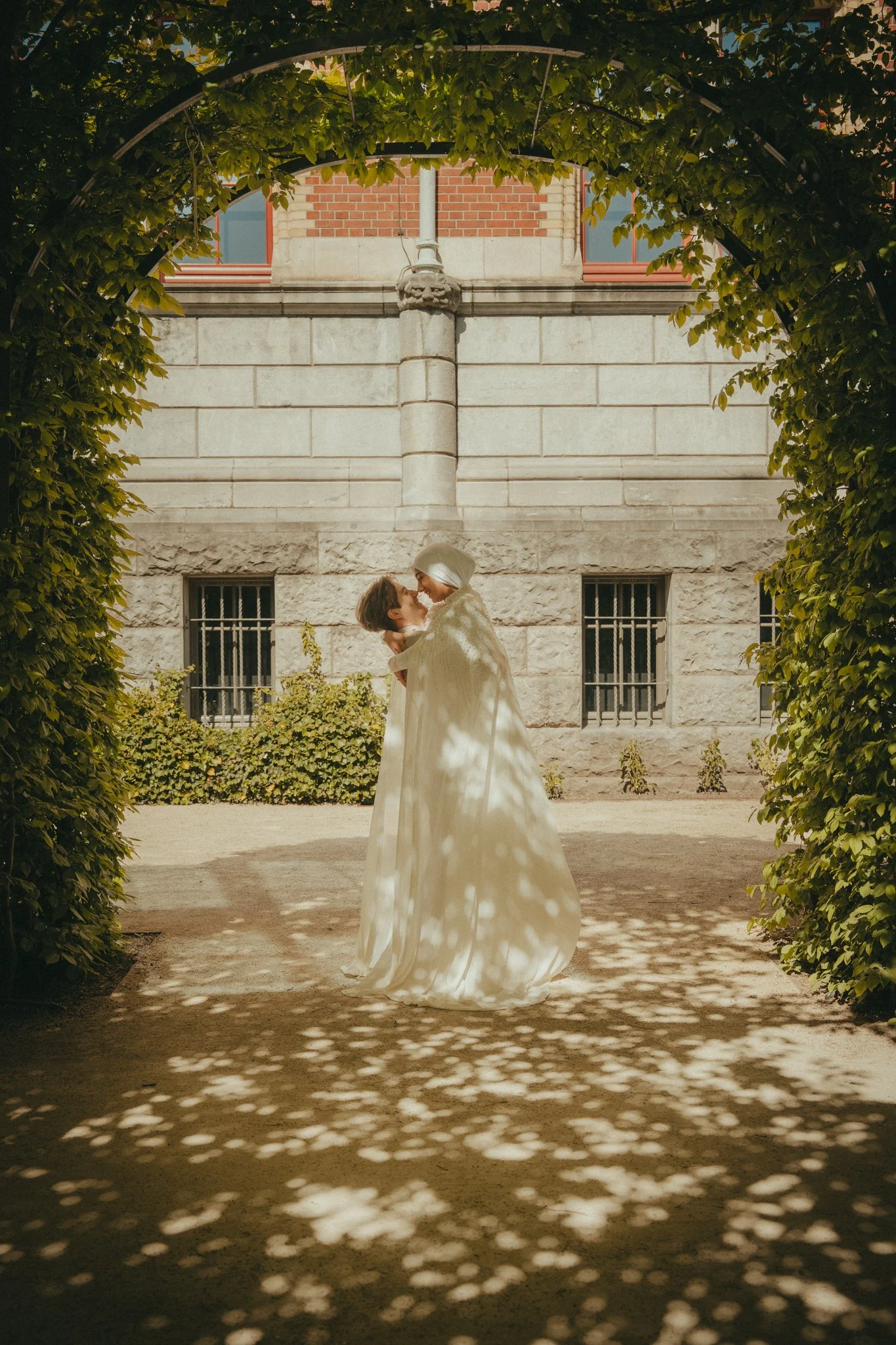 A couple dressed in wedding attire, the bride in a white gown and veil, dance closely under dappled sunlight created by tree leaves, in an outdoor garden with a historic stone building in the background.