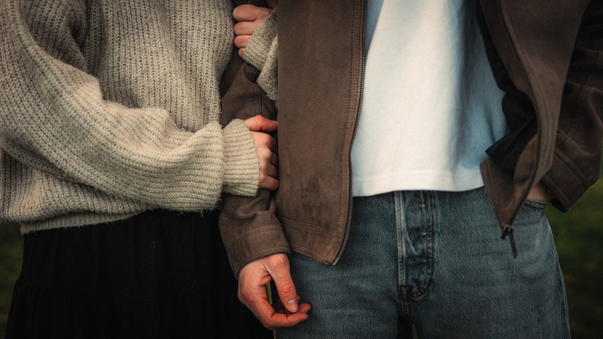 Two people are holding hands, with only their hands and part of their torsos visible. One person is wearing a beige sweater, and the other person is wearing a brown jacket over a white shirt and blue jeans.