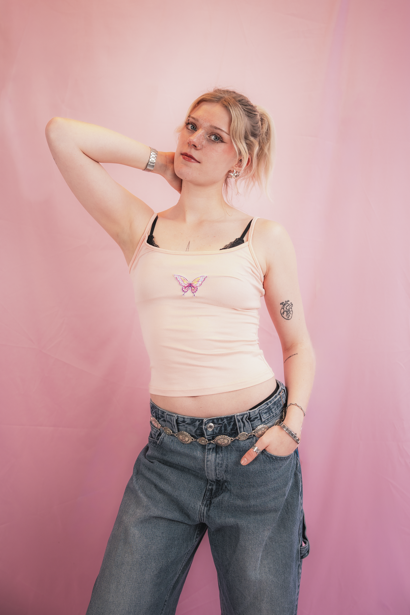 A young woman with blonde hair tied in a high ponytail, wearing a light pink tank top with a butterfly design, dark jeans with a decorative chain belt, and multiple rings and earrings. She has her right hand behind her head, standing against a pink b