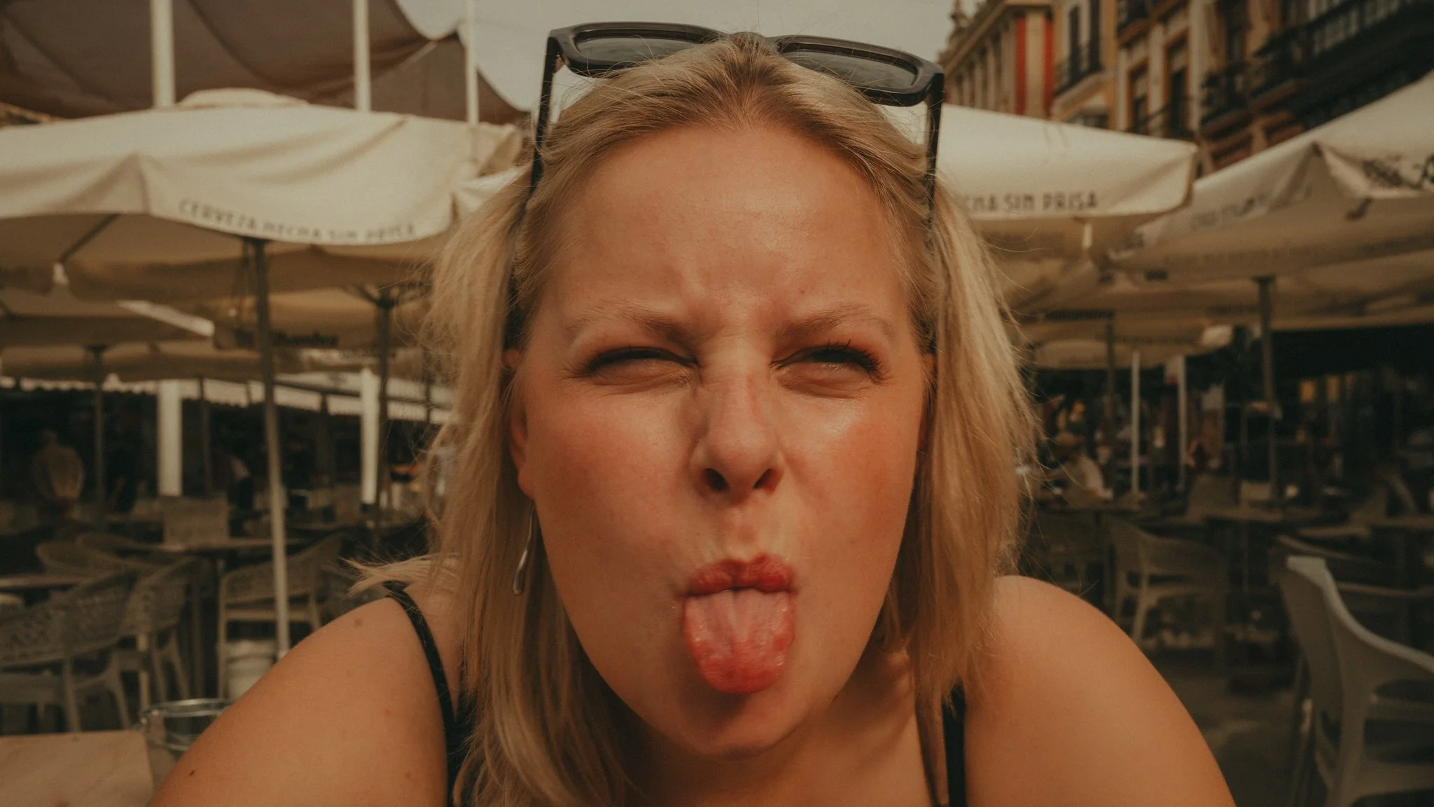 Close-up of a woman with long blond hair, wearing sunglasses on her head, making a silly face with her tongue out and lips puckered, sitting outdoors at a cafe or restaurant with umbrellas and buildings in the background.