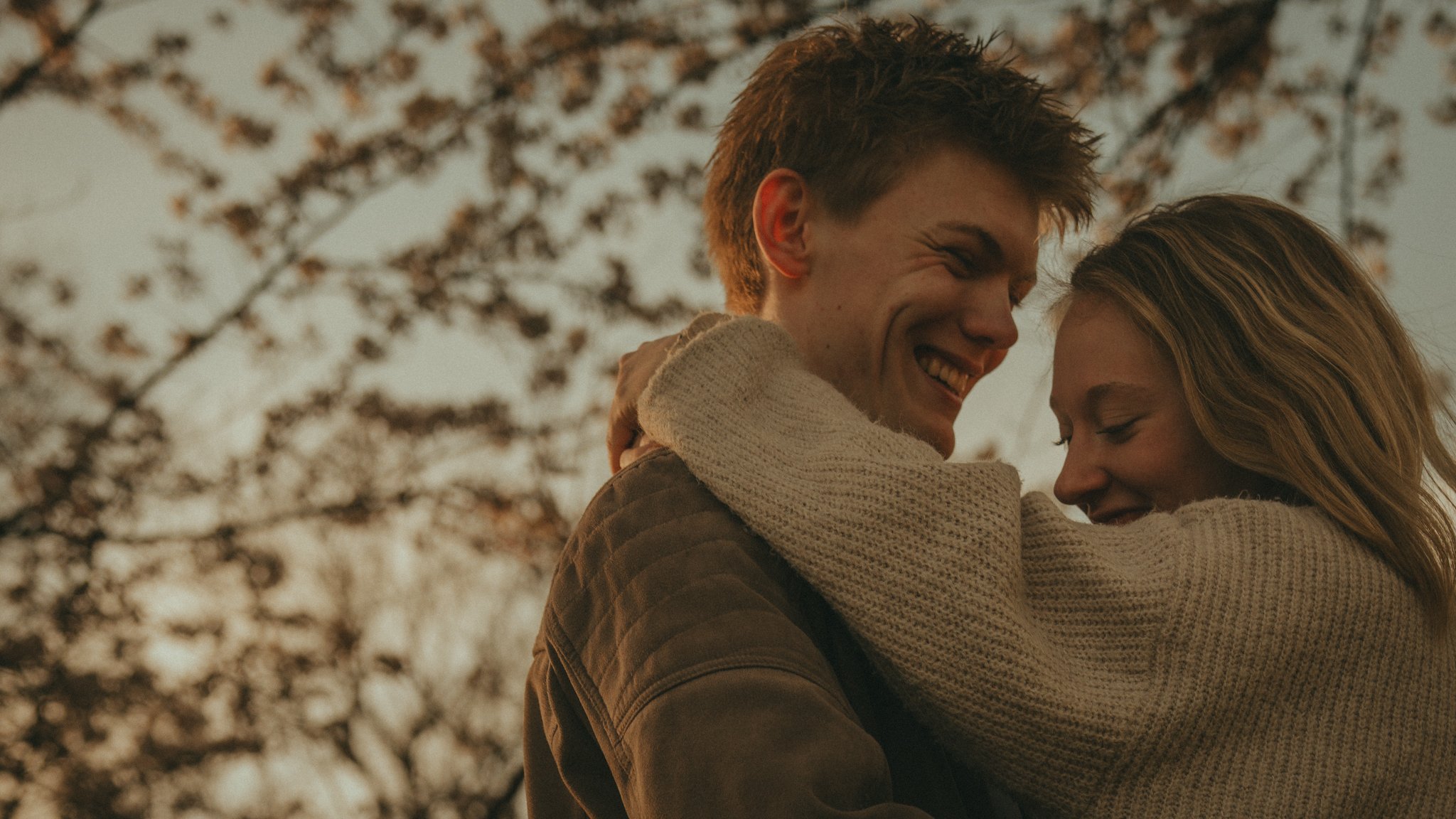 A young couple sharing a warm embrace outdoors, smiling and leaning close with trees in the background.