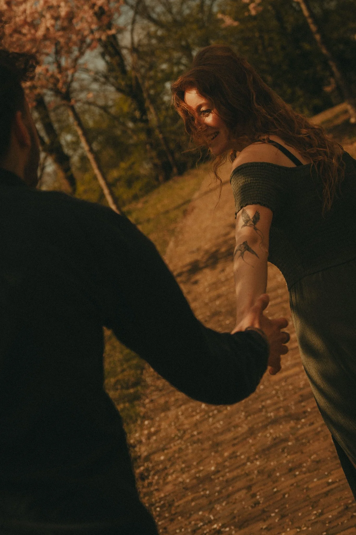 A woman with curly hair and tattoos on her arm is smiling and holding hands with a man during an outdoor scene at dusk or night, with trees and fallen leaves visible in the background.