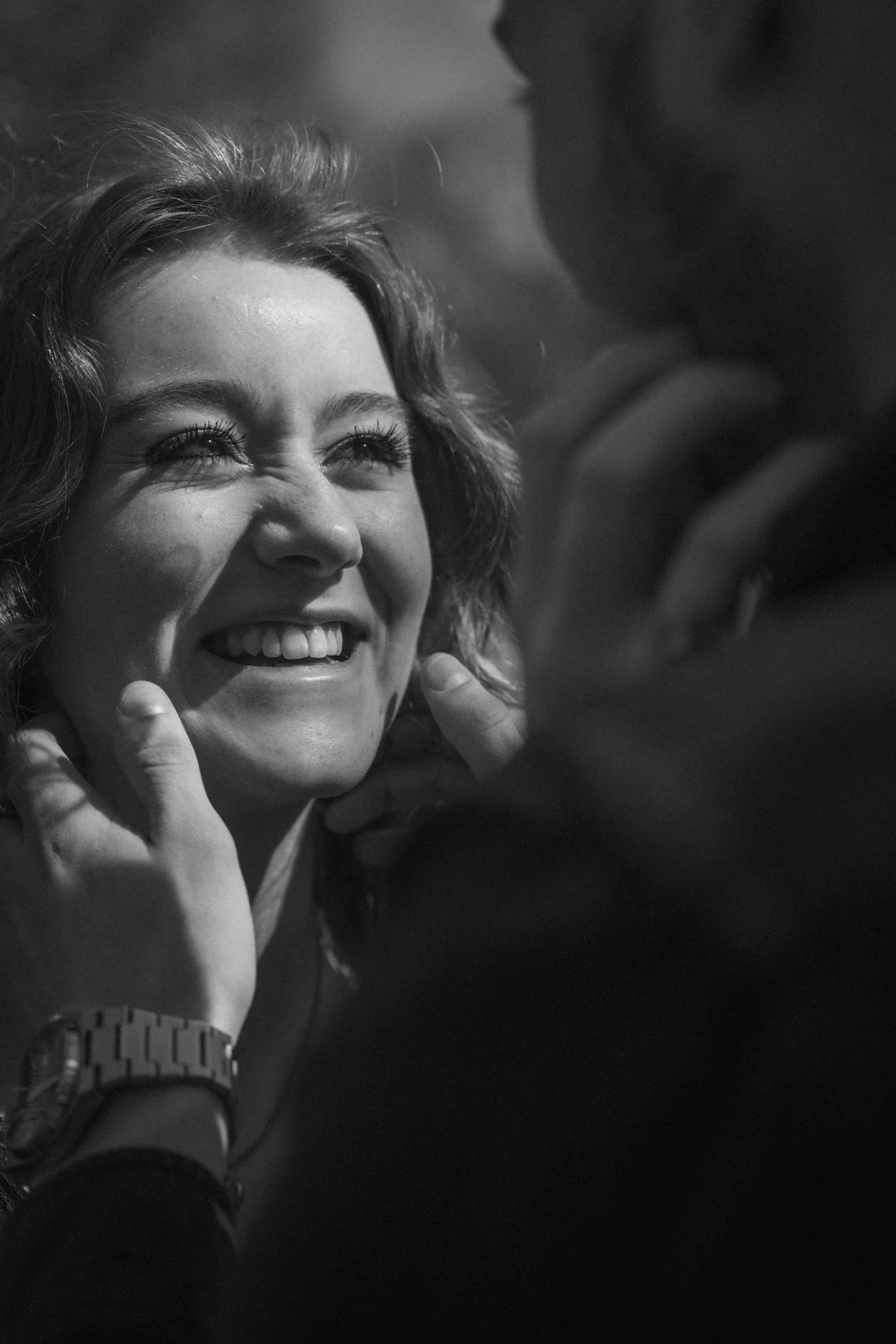 Black and white photo of a smiling woman with wavy hair, looking upwards, touching her face with one hand, wearing a watch.