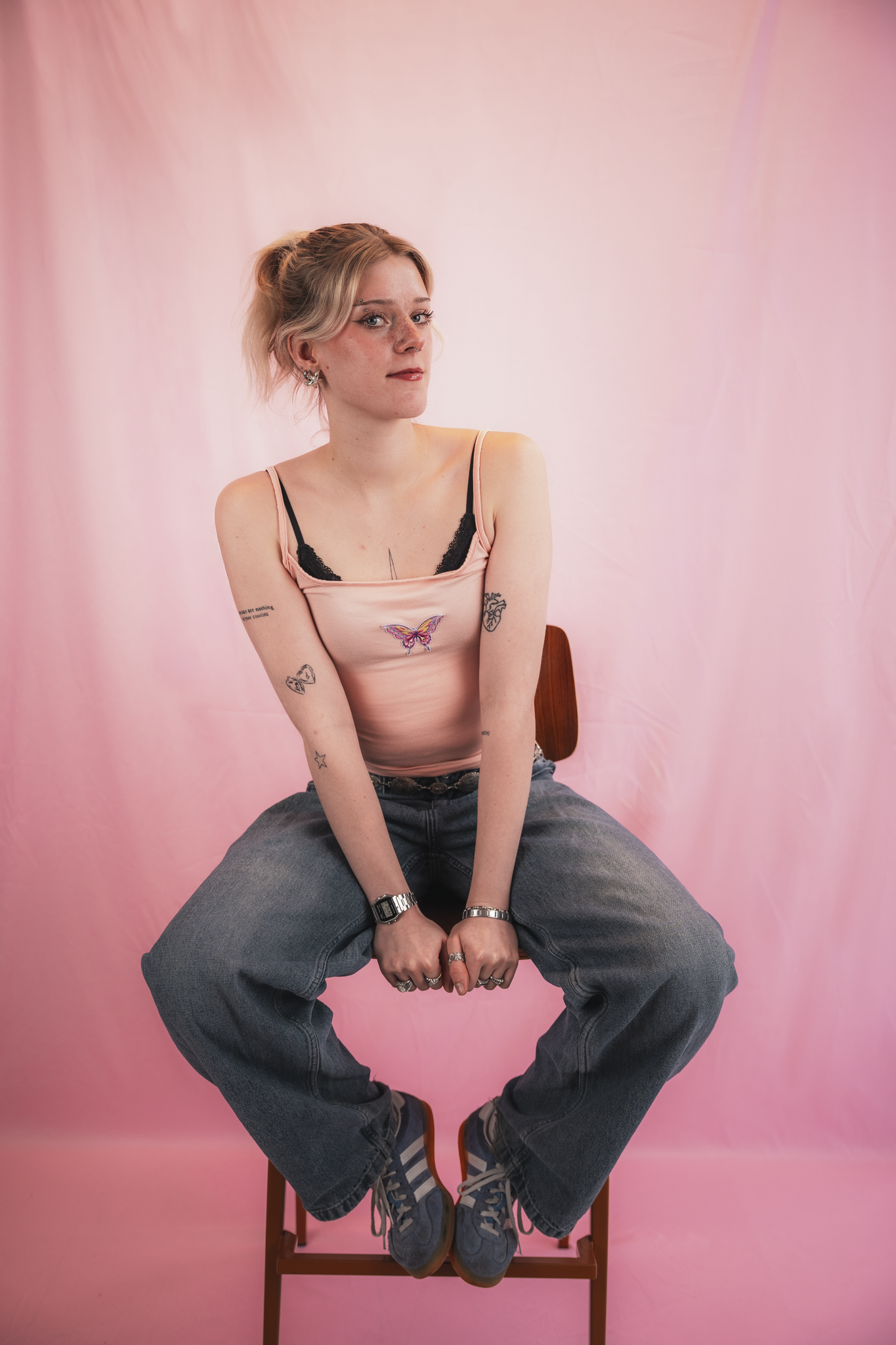 A young woman with short blond hair, tattoos on her arms, and ear piercings, sitting on a wooden chair against a pink backdrop. She is wearing a pink tank top with a butterfly design, loose jeans, and blue sneakers, with a relaxed expression and her 
