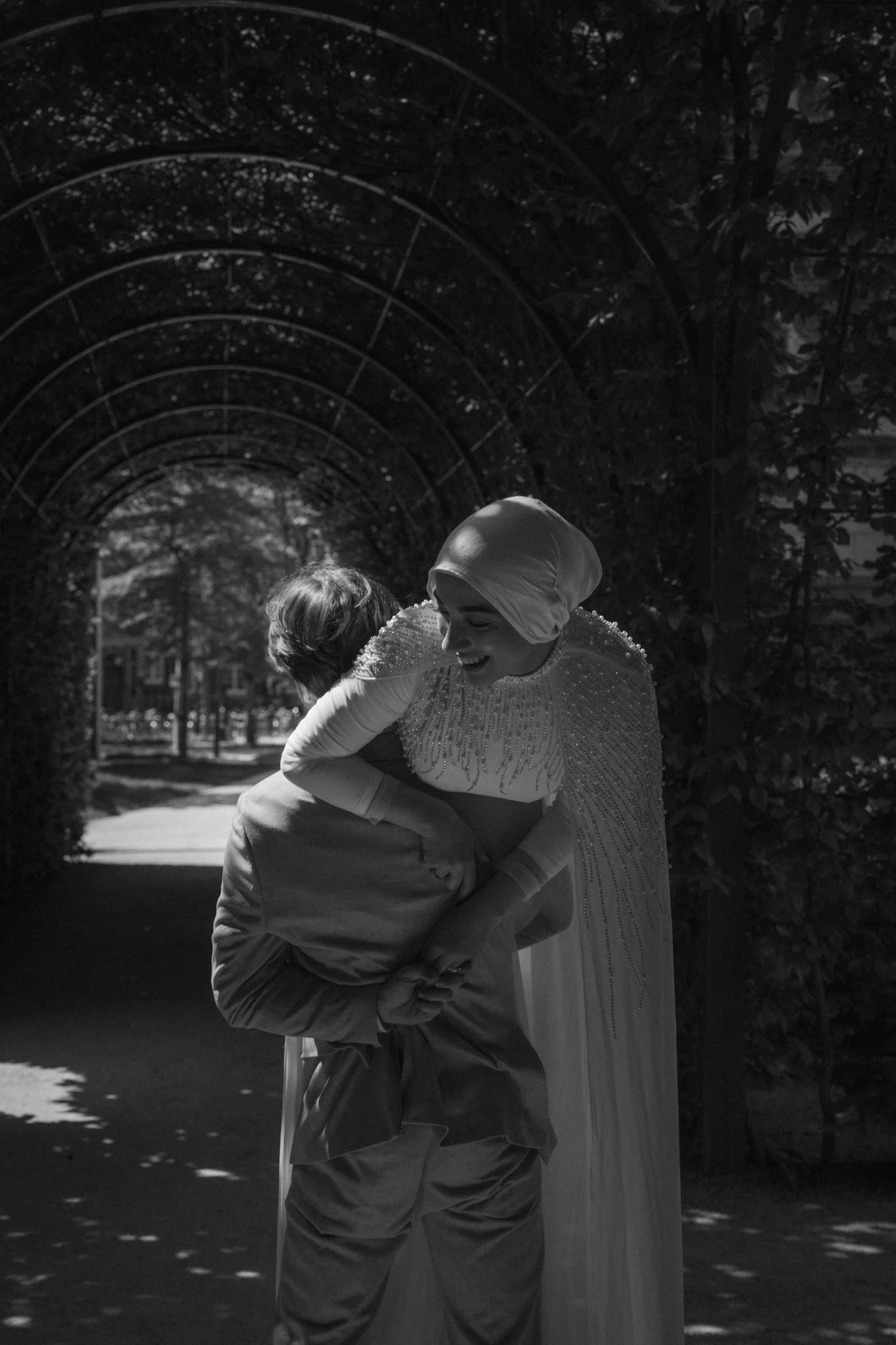 A joyful woman dressed in a fancy gown, wearing a head wrap, is hugging and lifting a young boy in a park during the daytime. The park has an arched trellis structure in the background and trees with sunlight filtering through.