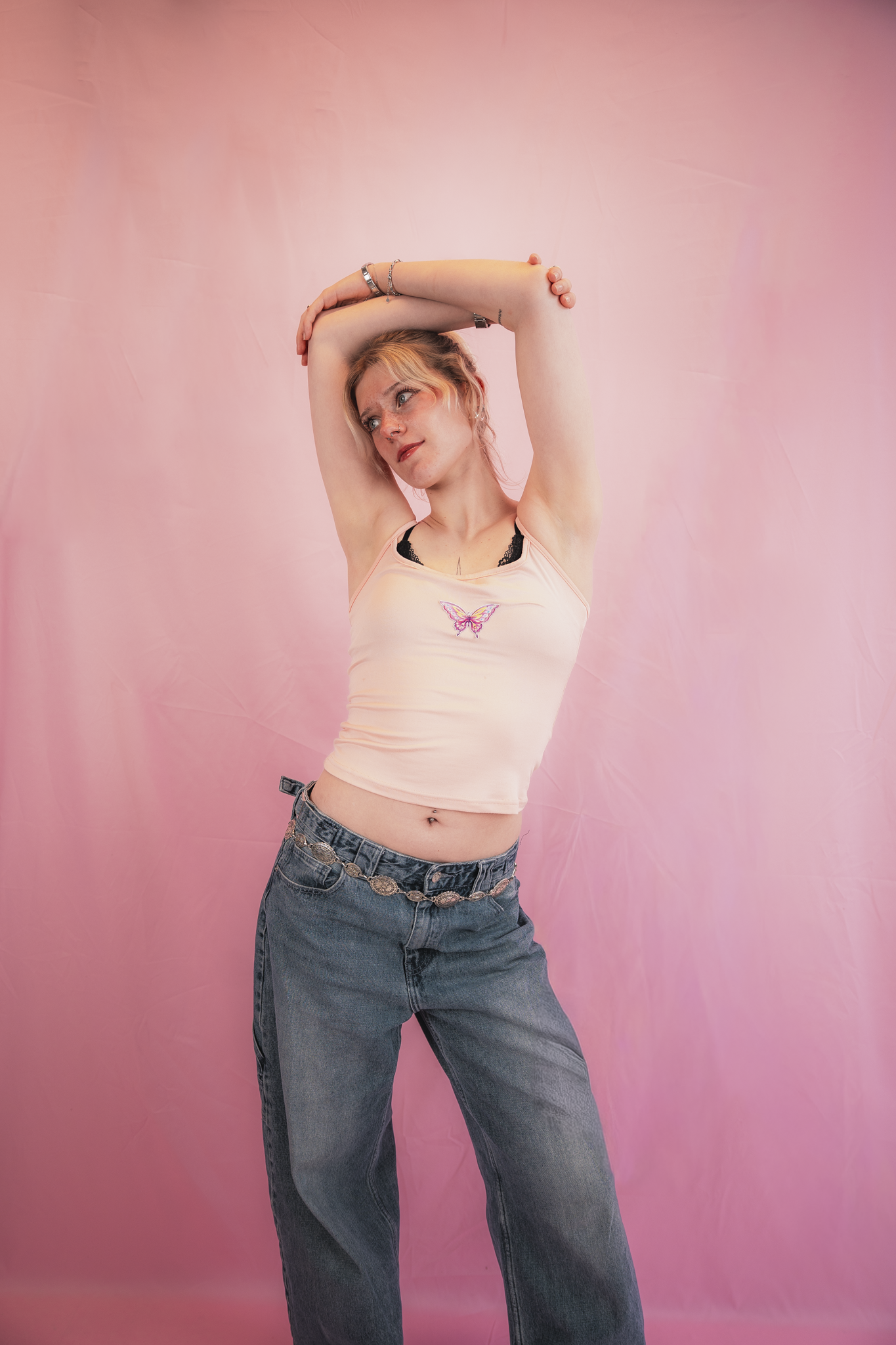 A young woman with blonde hair and fair skin is posing with her arms raised above her head against a pink backdrop. She is wearing a light pink tank top with a butterfly design and dark jeans, accessorized with bracelets and a chain belt.
