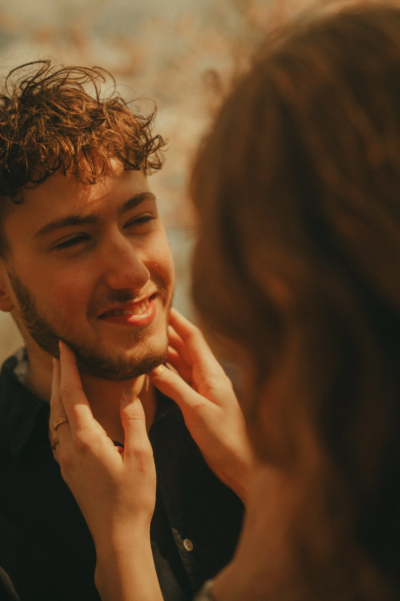 A young man with curly hair and a beard smiling while a woman touches his face.