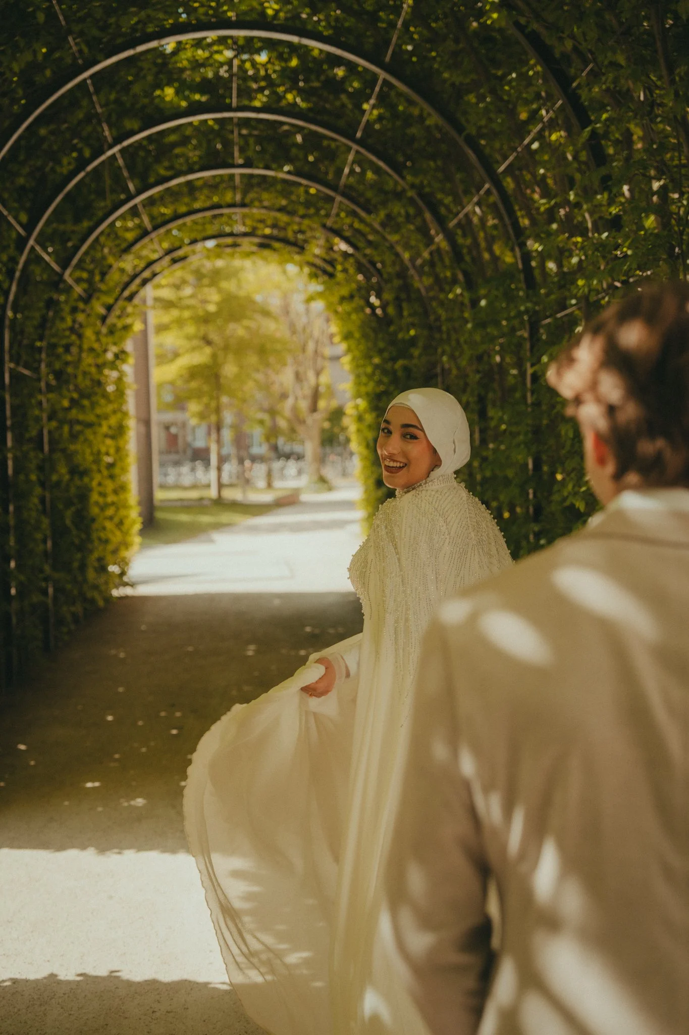 Two women wearing white dresses and headscarves under a green, arched tunnel with trees and sunlight in the background.