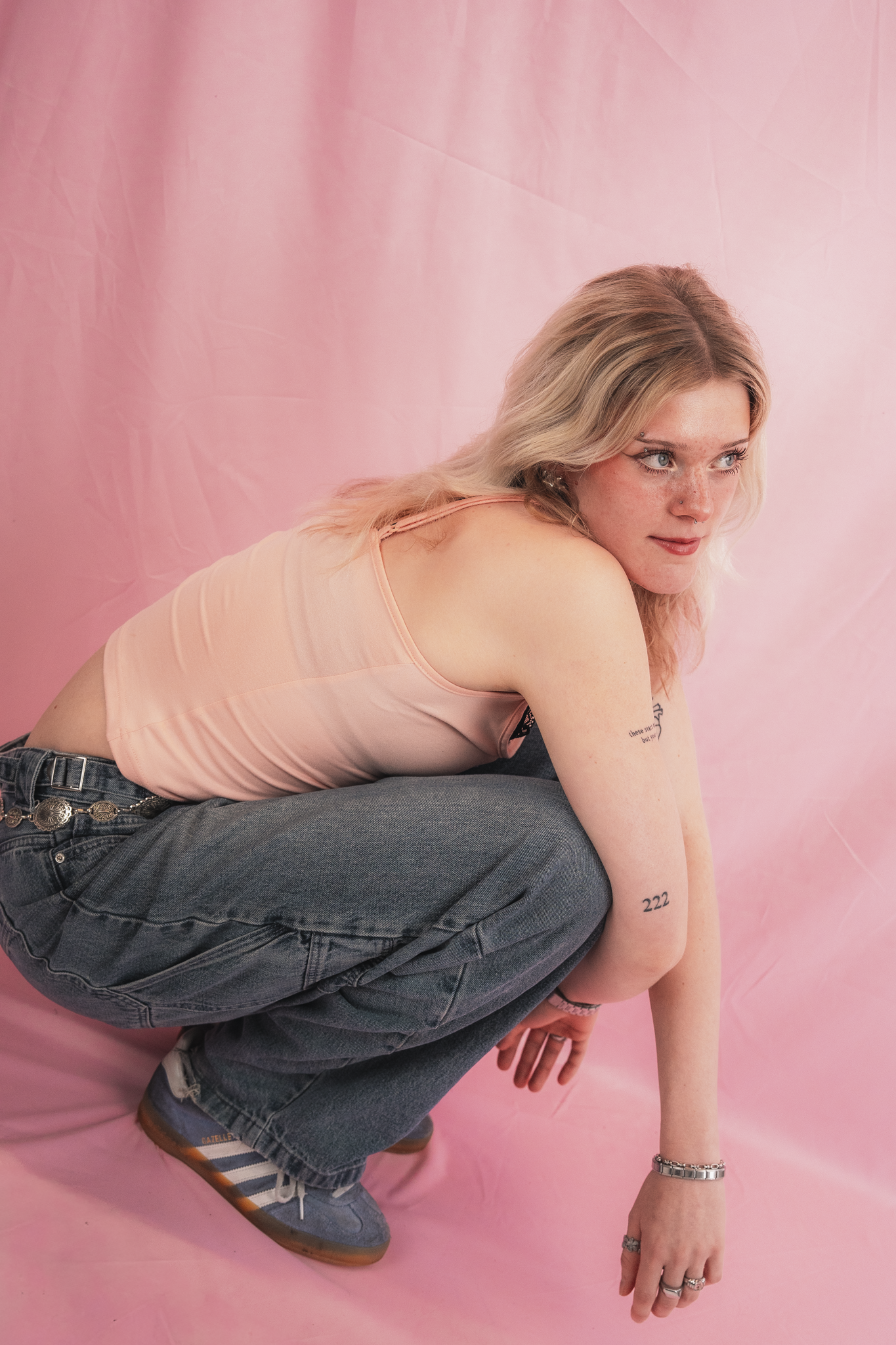 A woman with blonde hair and blue eyes posing against a pink backdrop, wearing a peach-colored tank top, blue jeans with a belt, and sneakers, with tattoos on her arm and tattoos on her neck.