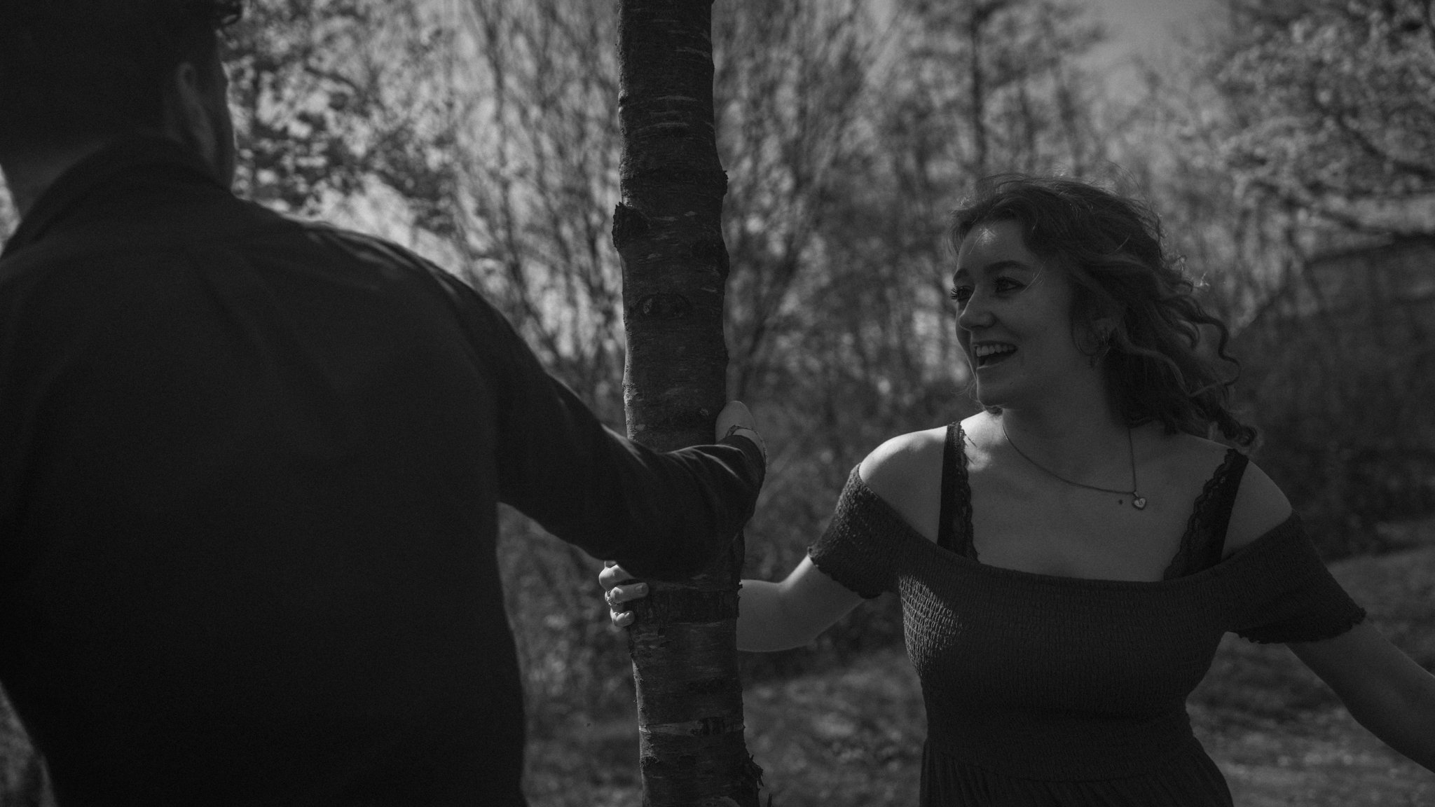 A black and white photo of a woman smiling, facing a man with his back to the camera, outdoors near a tree, with the woman holding the man's arm.