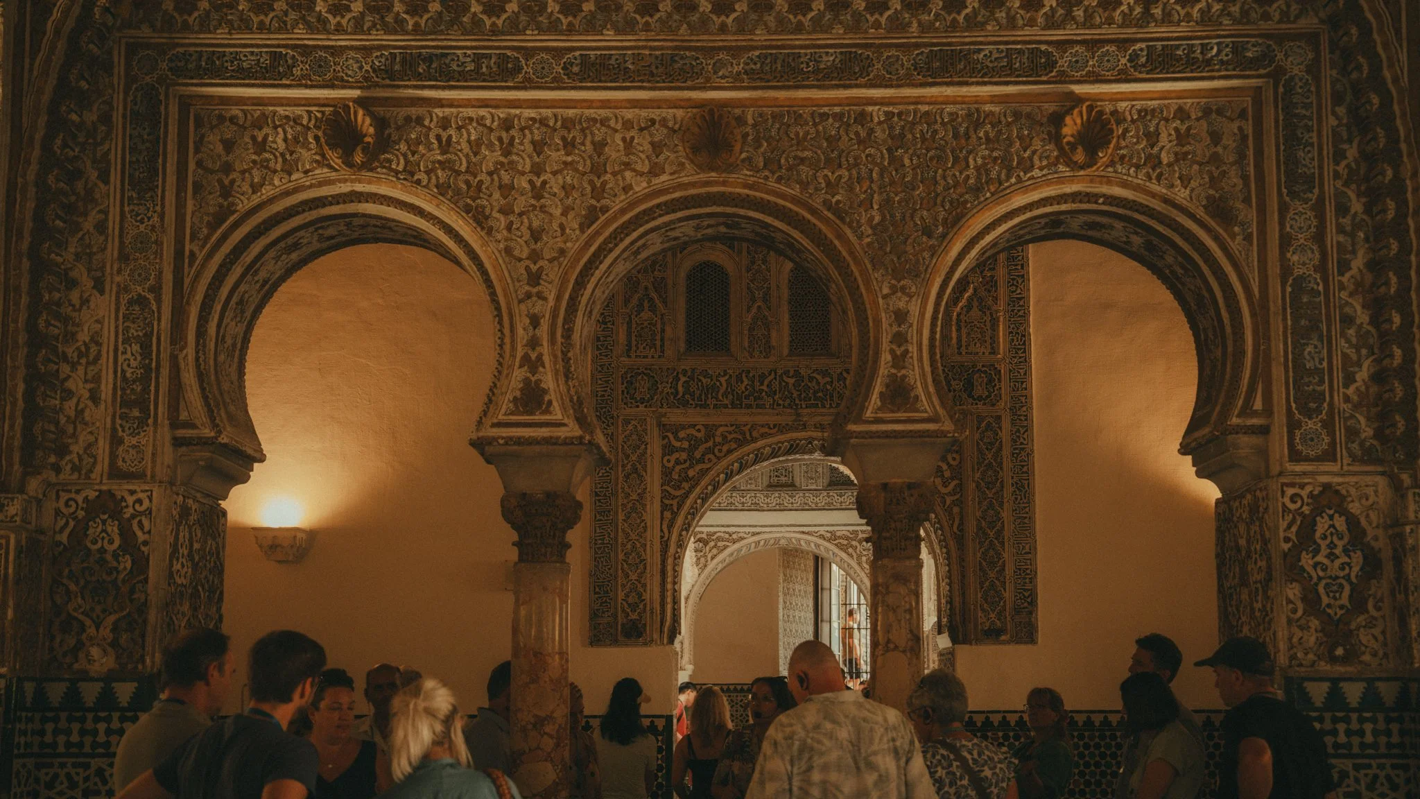 Interior view of a historic building with ornate, patterned arches and niches, filled with people seated and standing, with natural light coming through windows.