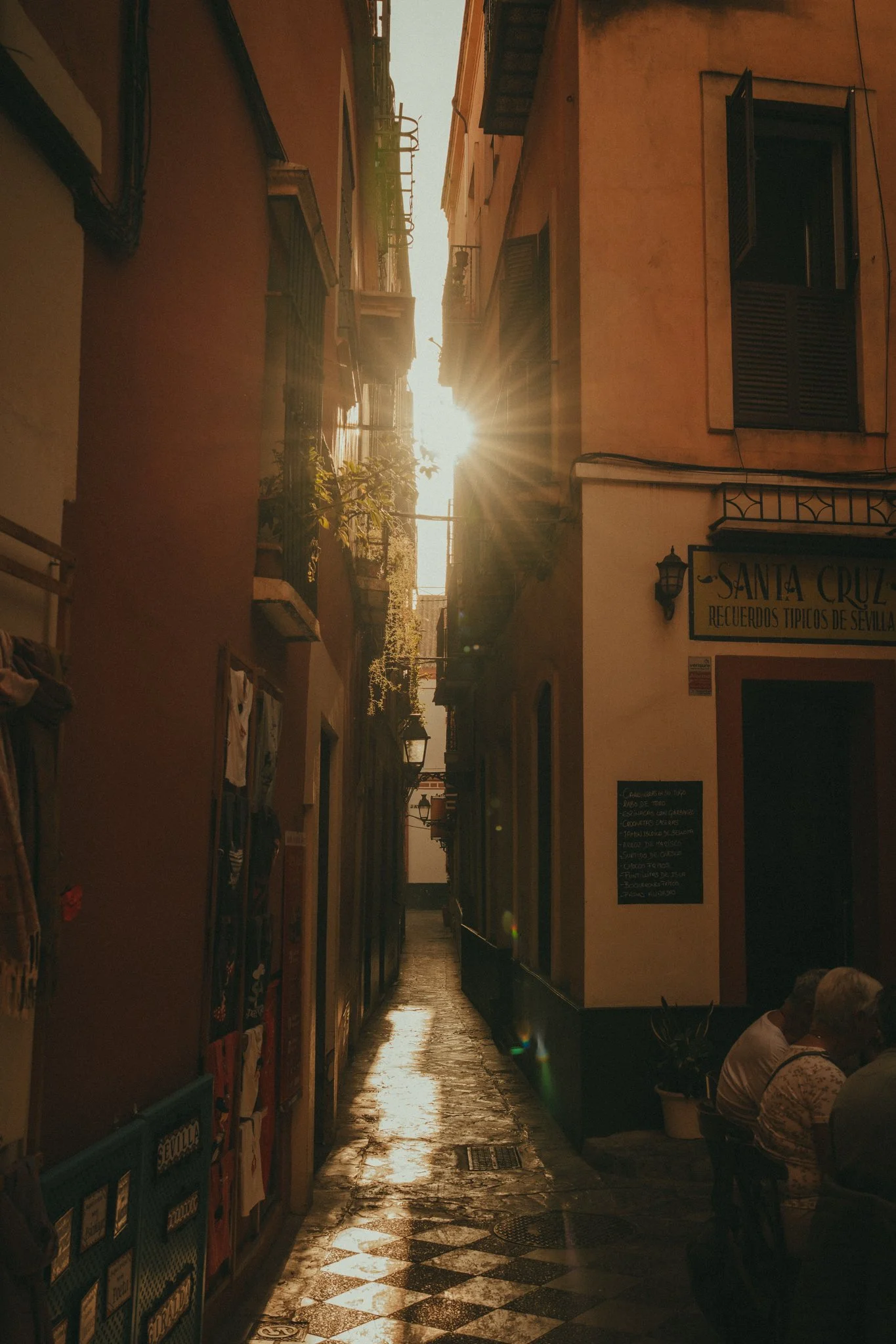 Narrow street with cobblestone pavement and buildings on either side, sunlight shining through at sunset, with an outdoor seating area and a sign for a business named Santa Cruz.