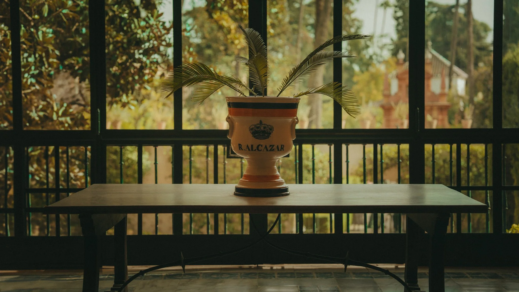 A decorative vase with palm leaves sits on a wooden table near a window with a view of trees and a building in the background.