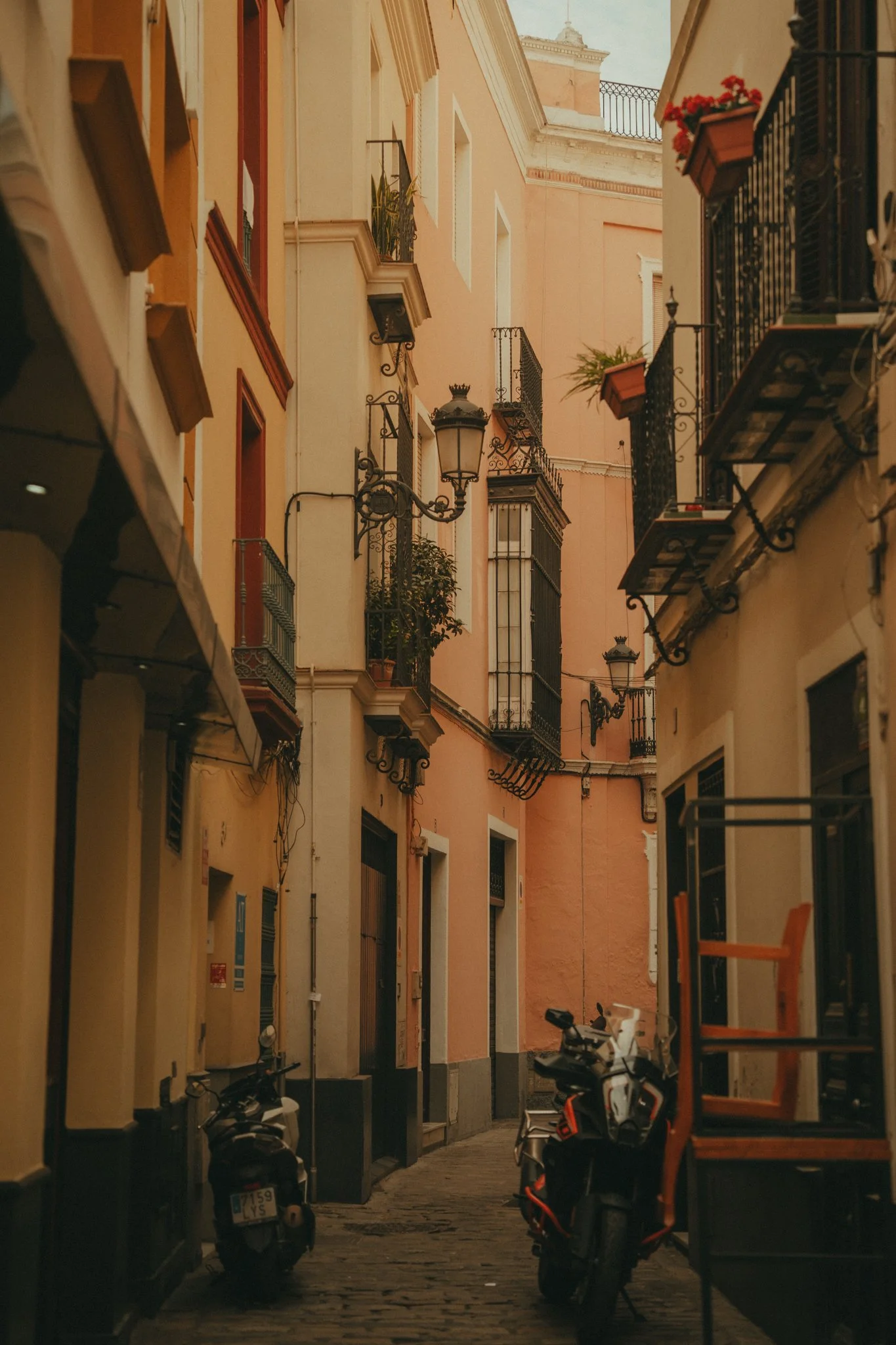 A narrow European street with pastel-colored buildings, balconies with potted plants, street lamps, and parked scooters.