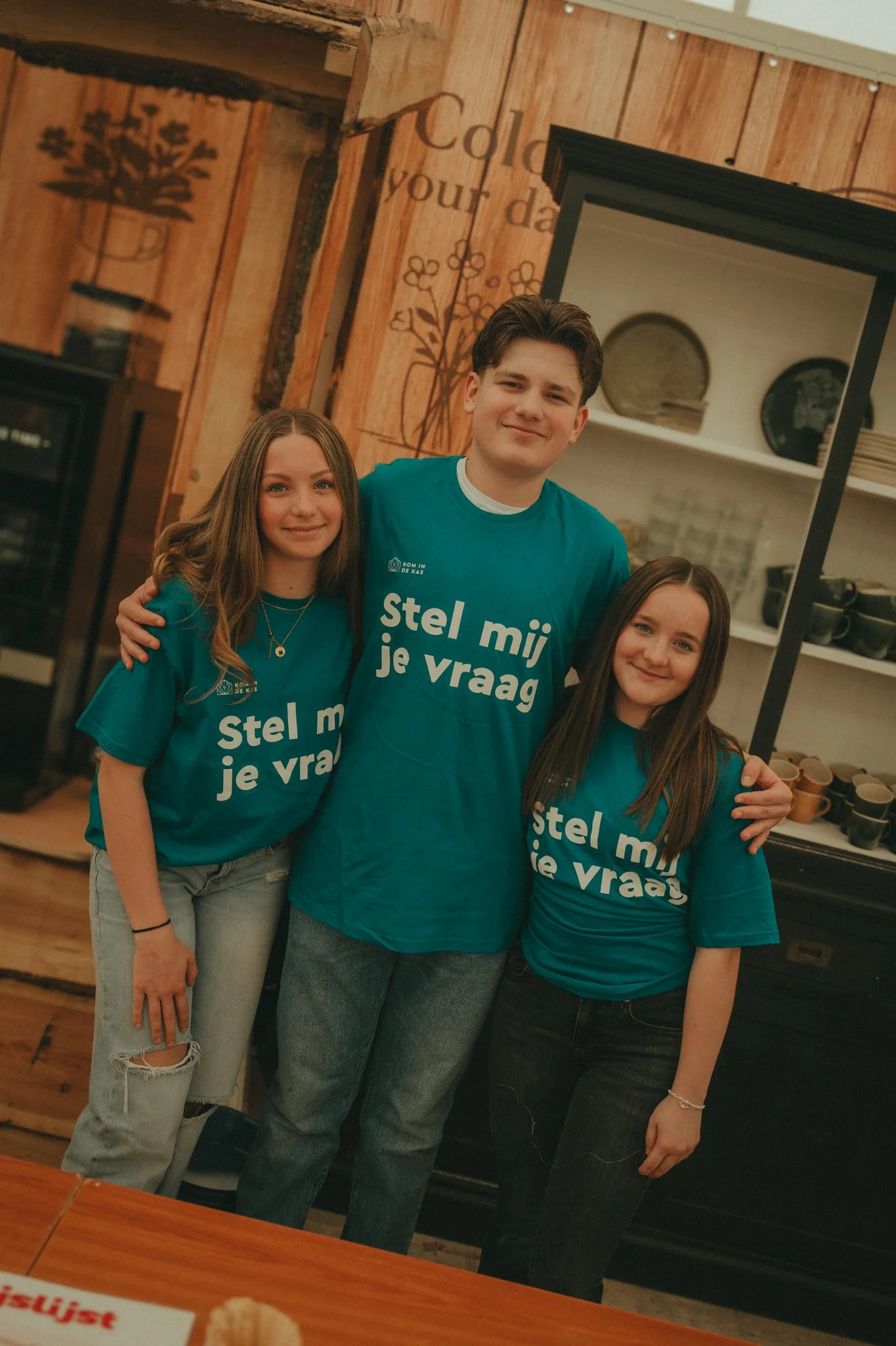 Three young people standing together, smiling in a rustic wooden interior, wearing matching teal T-shirts with Dutch text that says 'stel mij je vraag'.